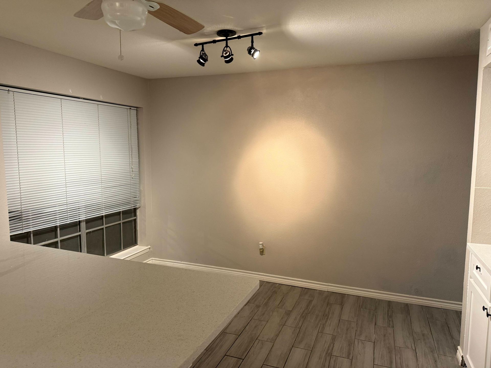 Empty room with window and track lighting; off-white walls, white blinds, and light gray wood-look flooring.
