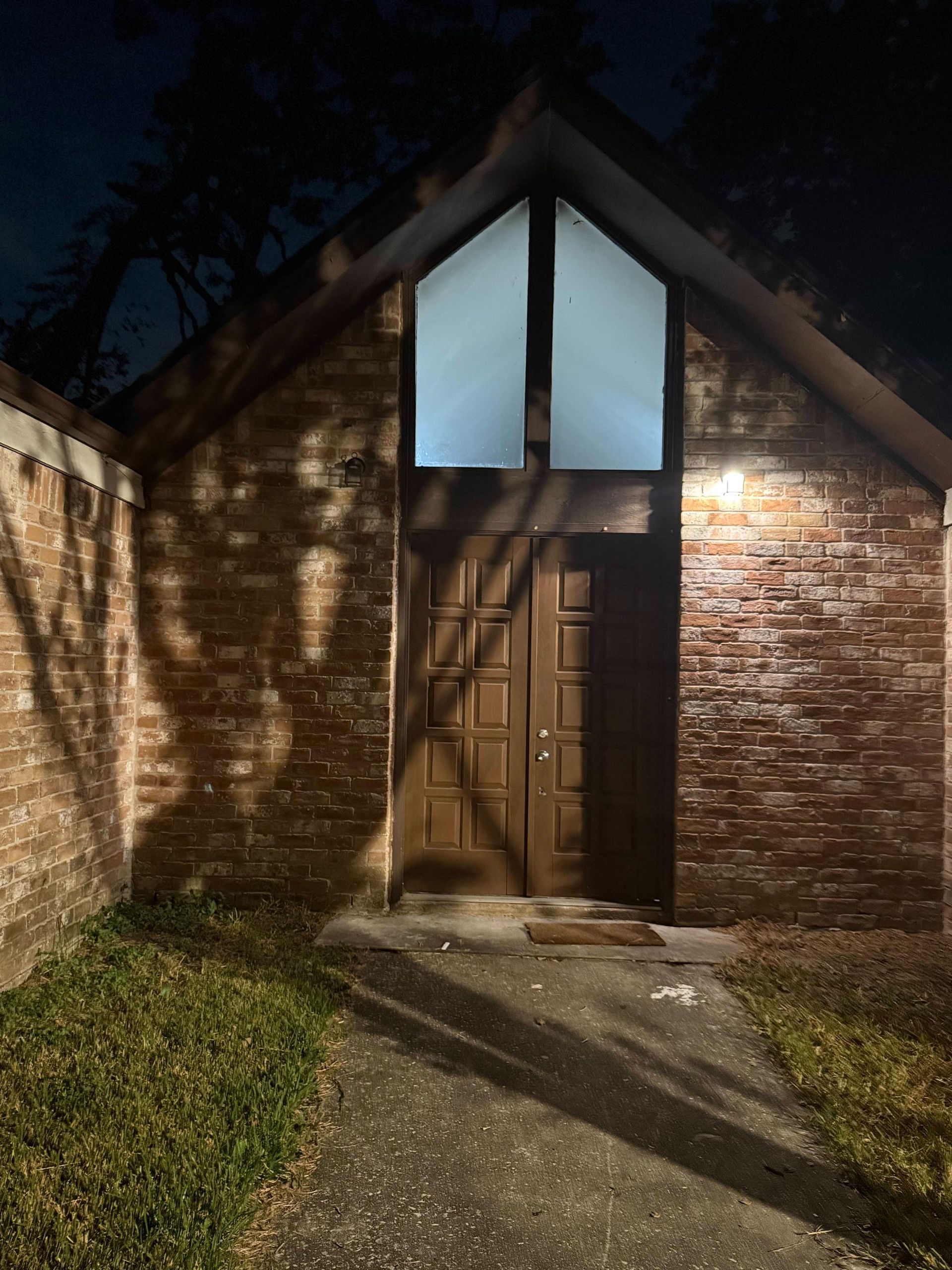 Brick building at night with a gabled roof. A doorway, lit window, and shadows are visible.