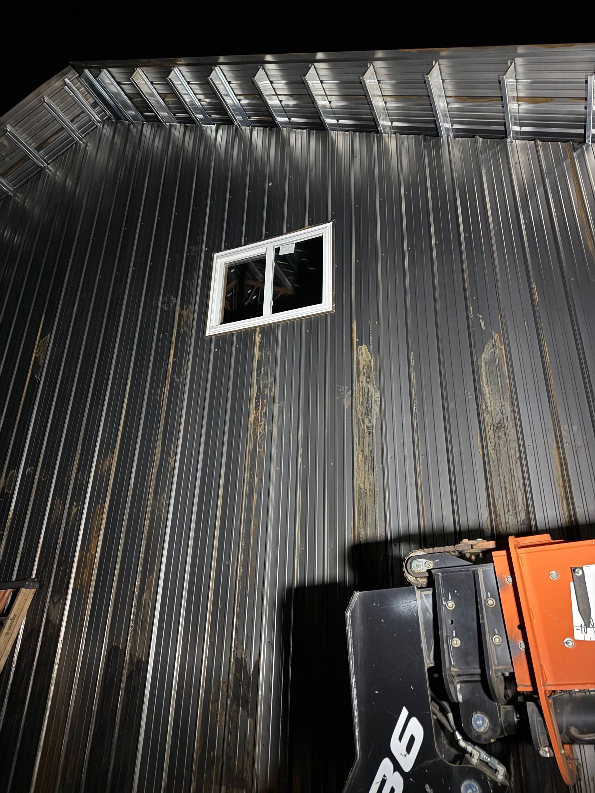 Metal building exterior with a small window, viewed from a low angle. An orange machine is visible in the lower right.