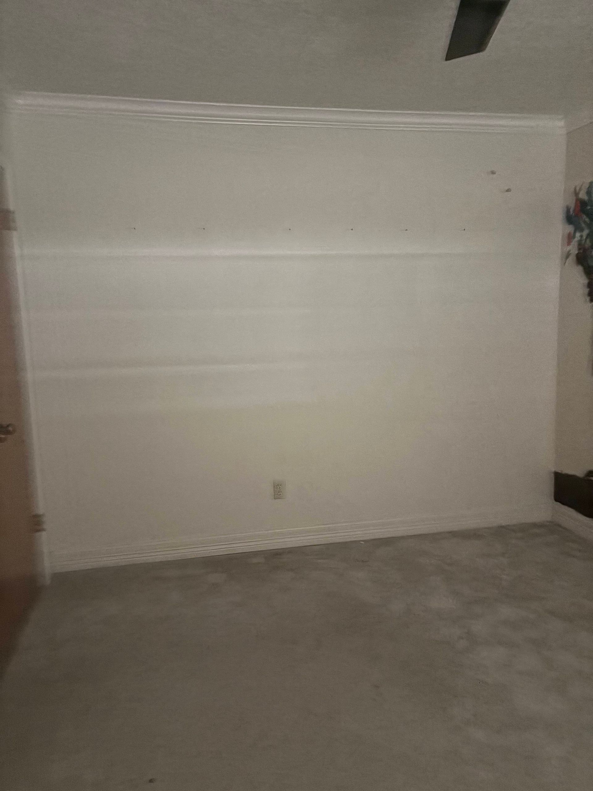 Empty, cream-colored room with a light gray carpet. Wall has crown molding and an electrical outlet.