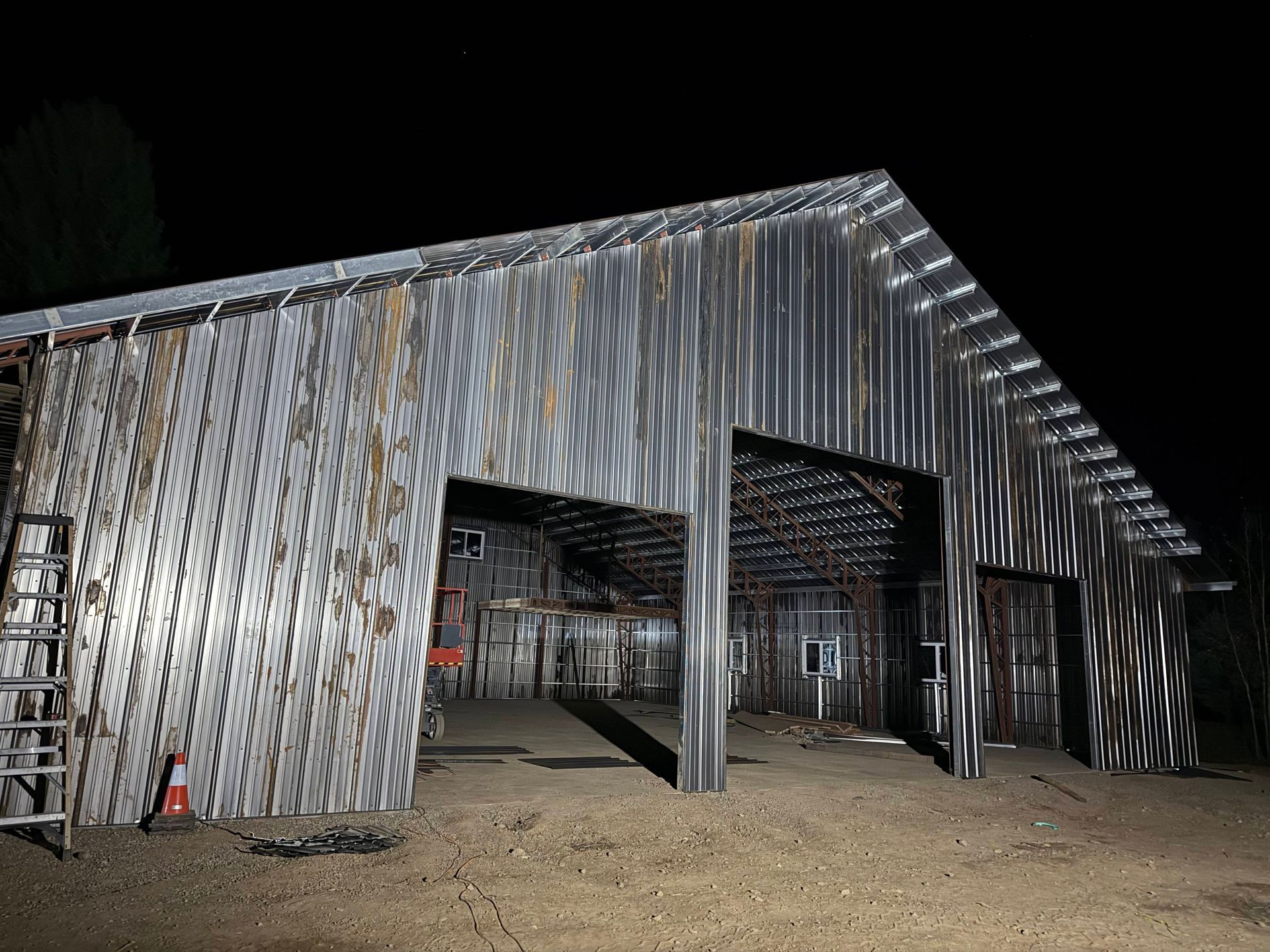 Metal-sided barn at night with open doors, illuminated interior, and a ladder on the side.