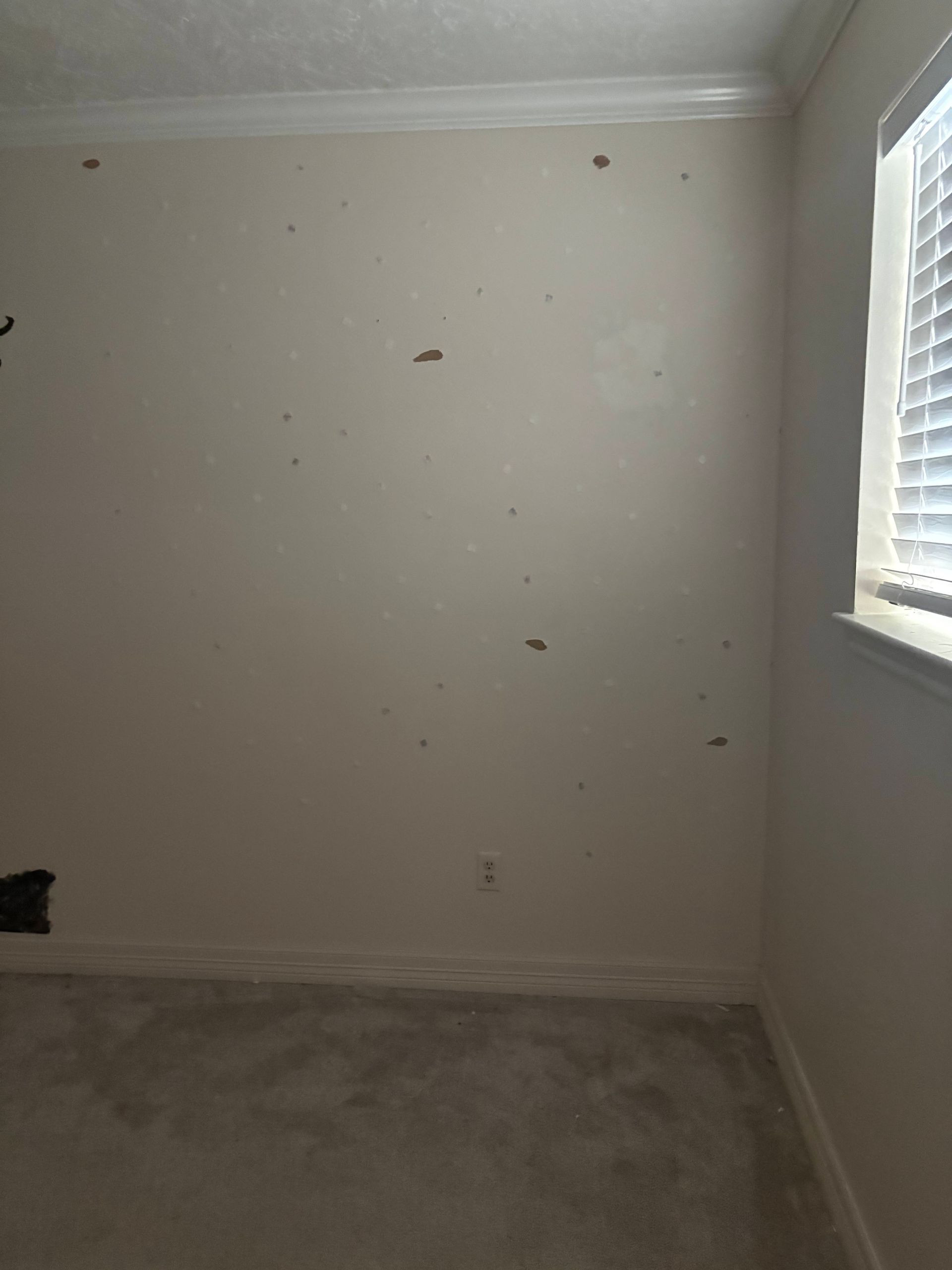 Bare interior wall with several holes and blemishes, next to a window with blinds.
