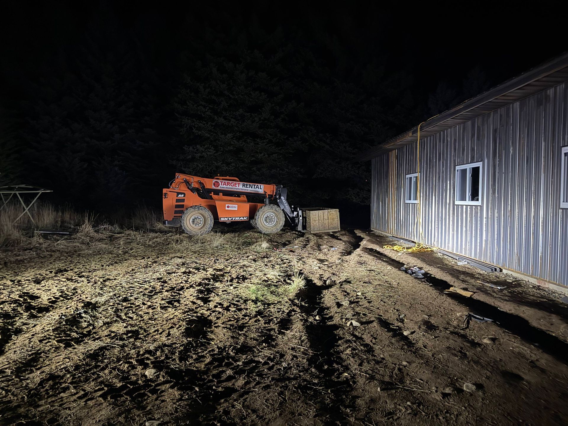 Orange telehandler moving a bale of hay near a metal barn at night.