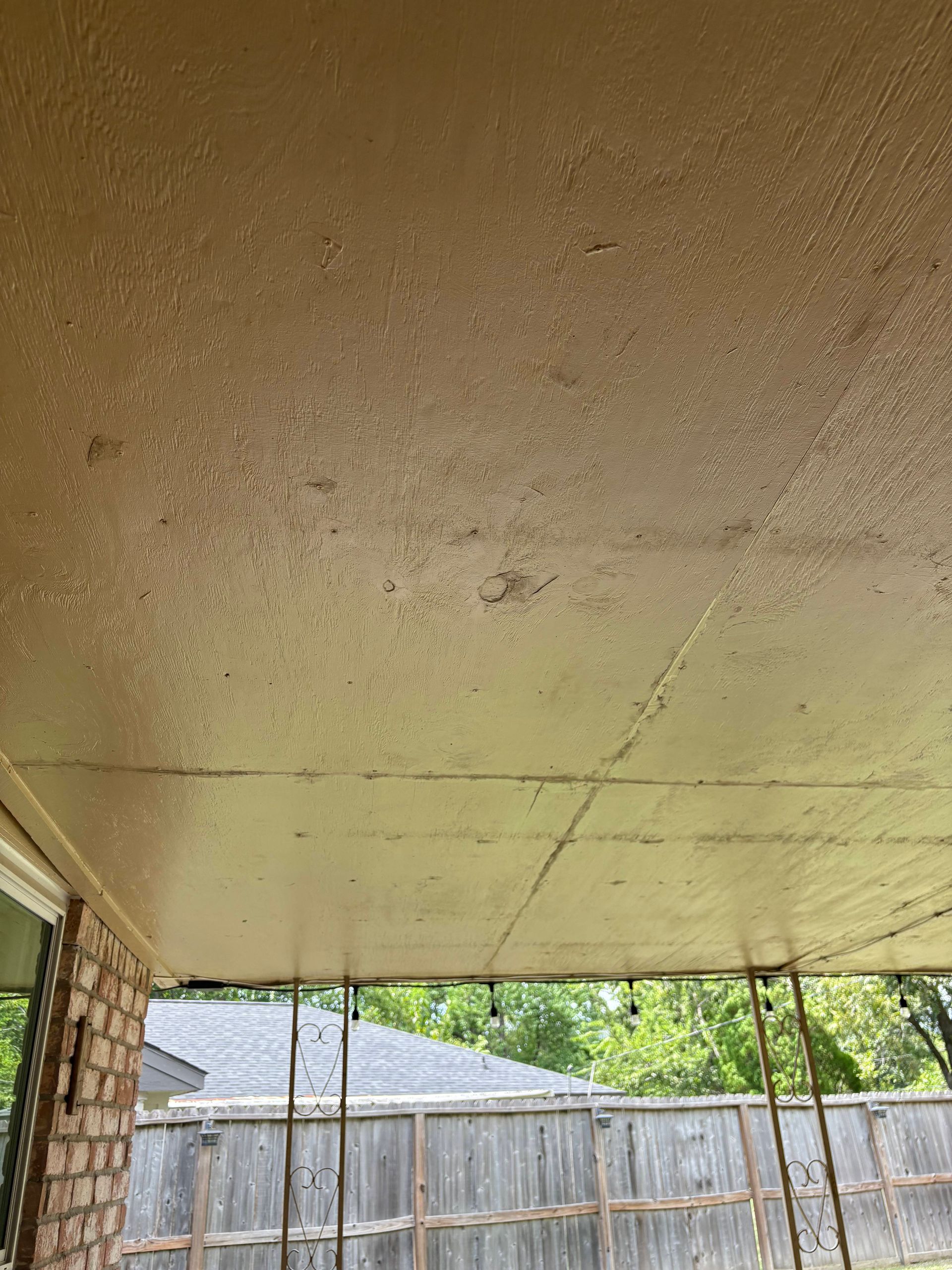 Beige painted ceiling of an outdoor patio with a wooden fence and trees in the background.