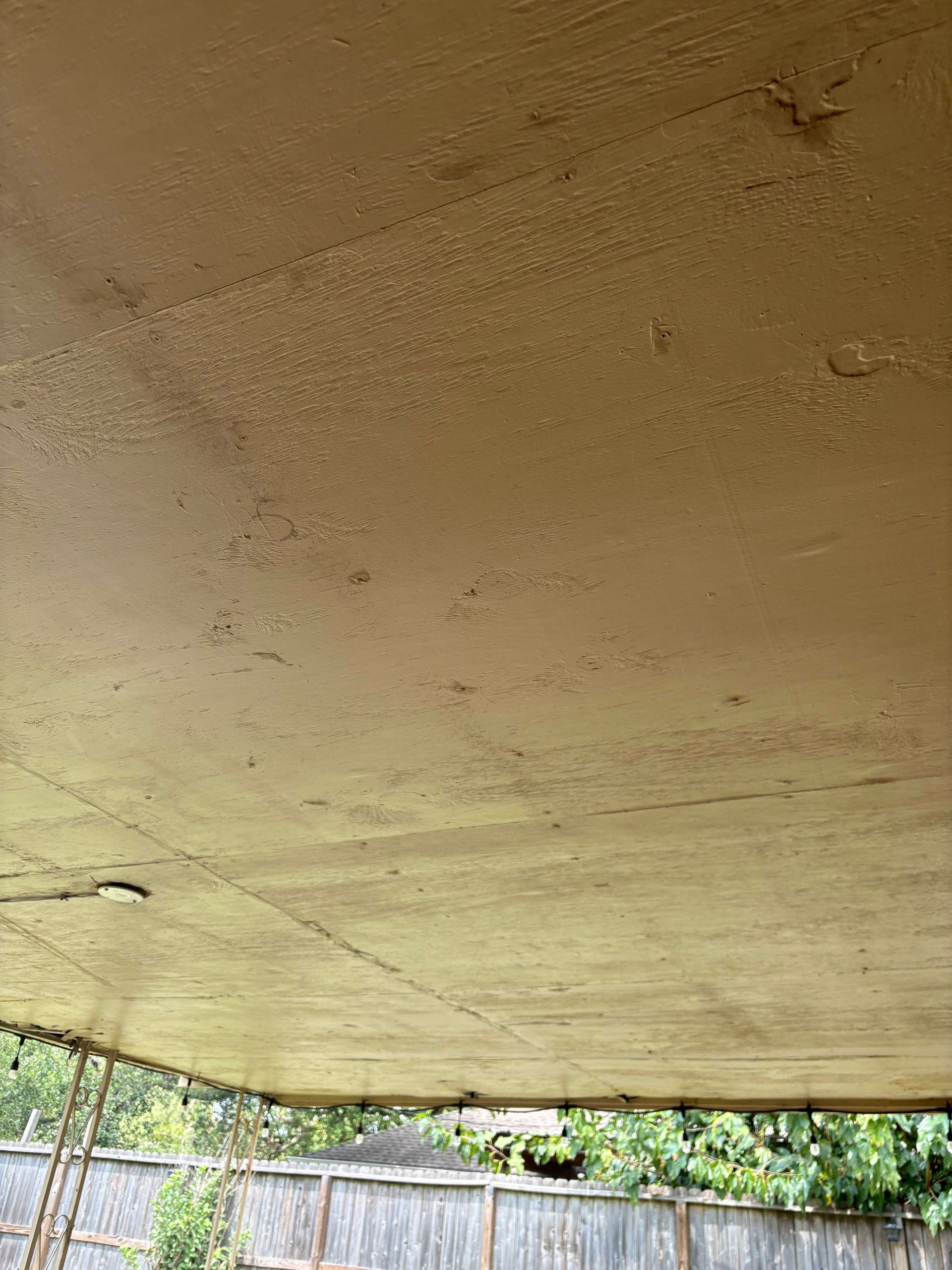A tan-colored, outdoor ceiling with visible imperfections, facing a green yard and wooden fence.