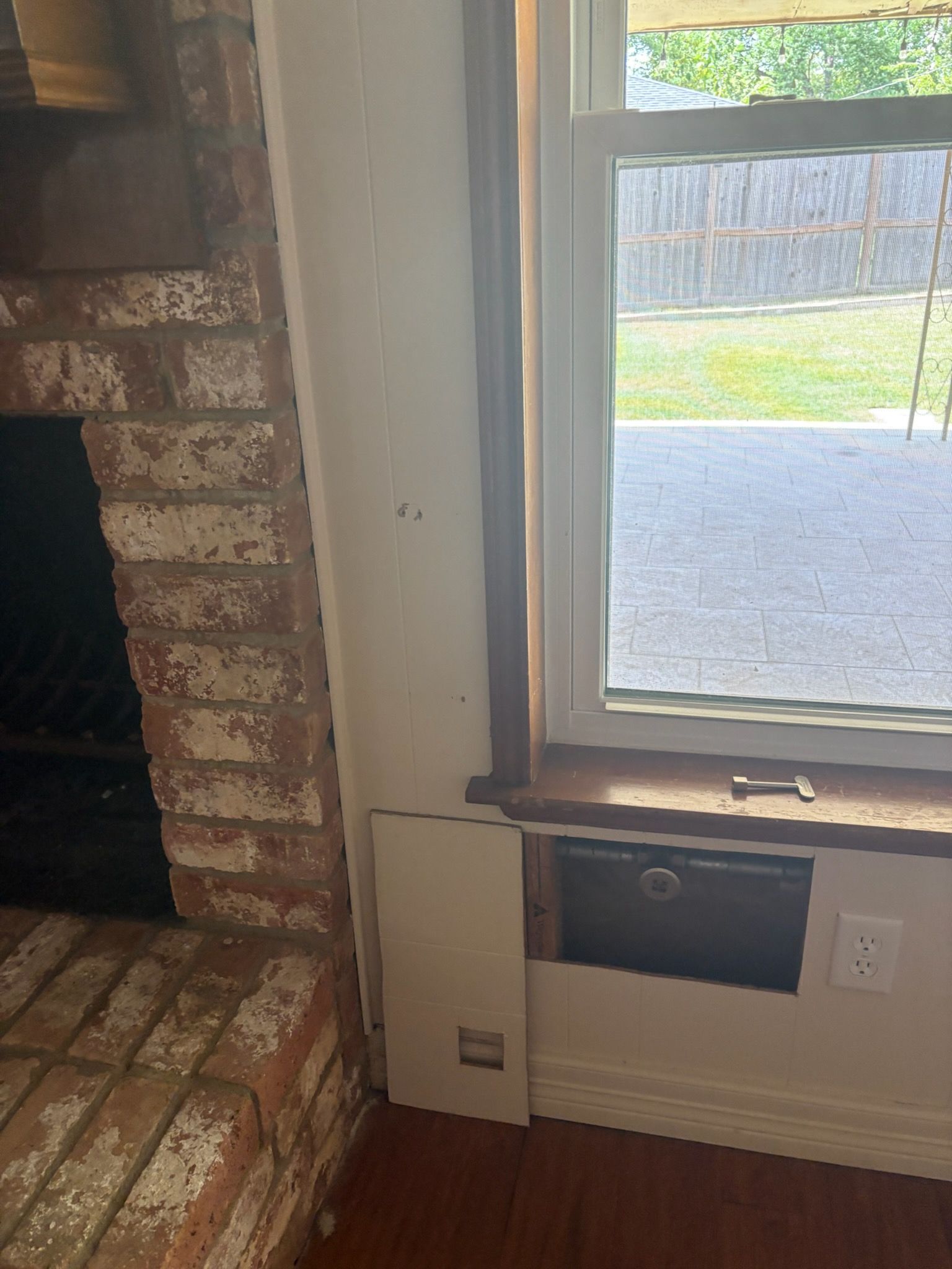 A brick fireplace and window with a wall vent and open access panel in a room.