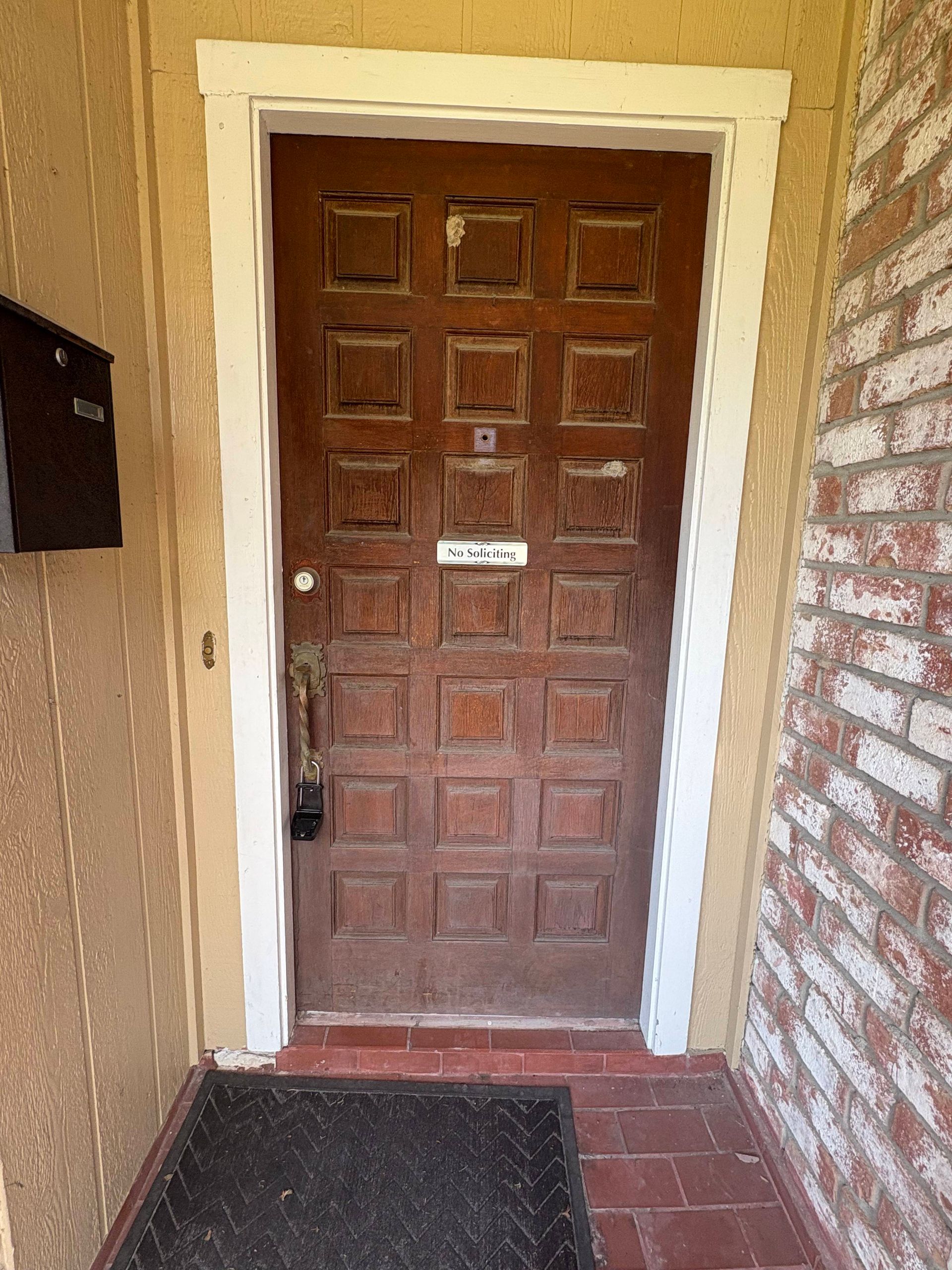 Brown, square-paneled door with white trim, a black mailbox, and brick wall on the right.