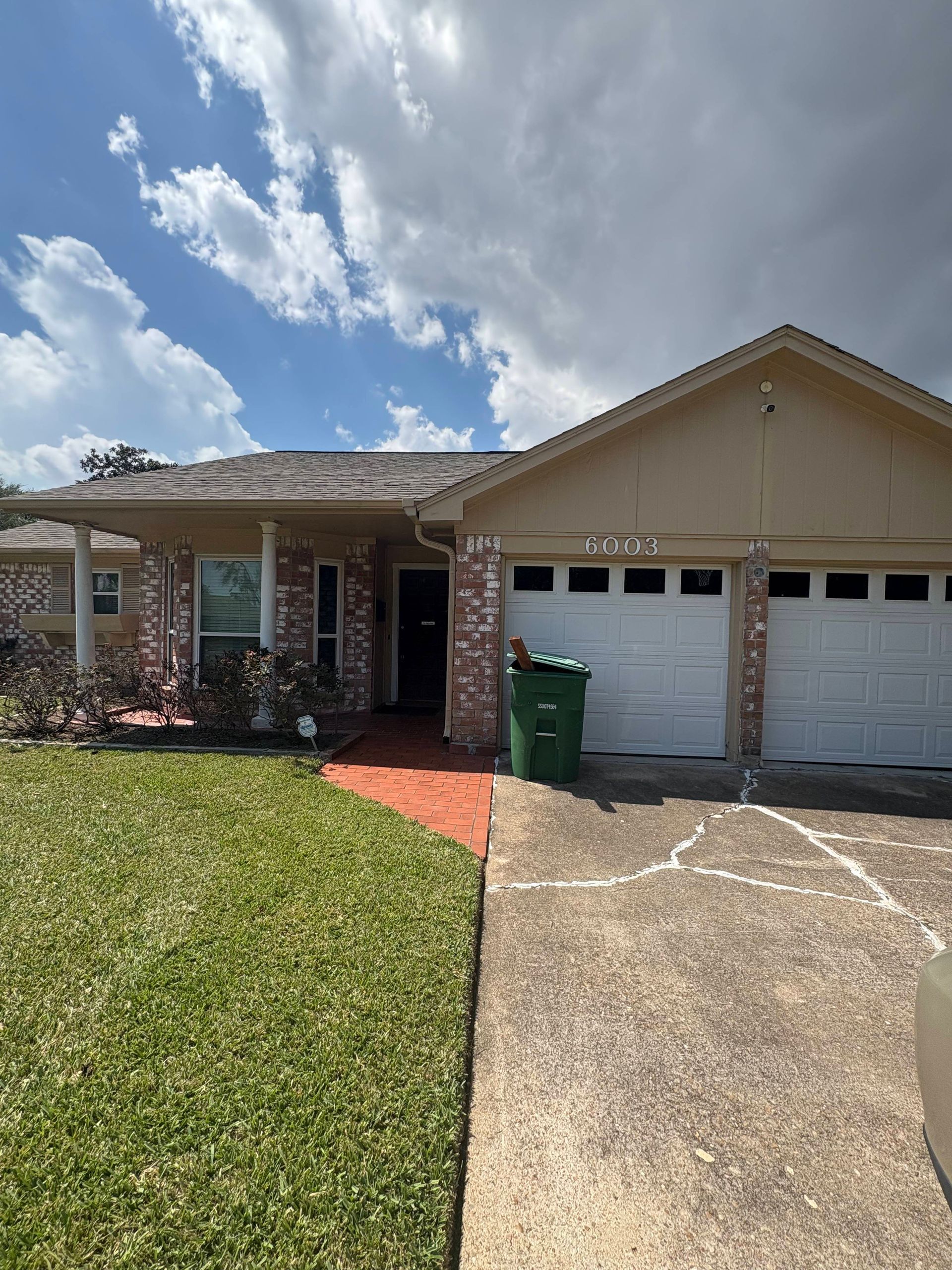 Tan brick house with two-car garage, green bin, and small front lawn on a cloudy day.