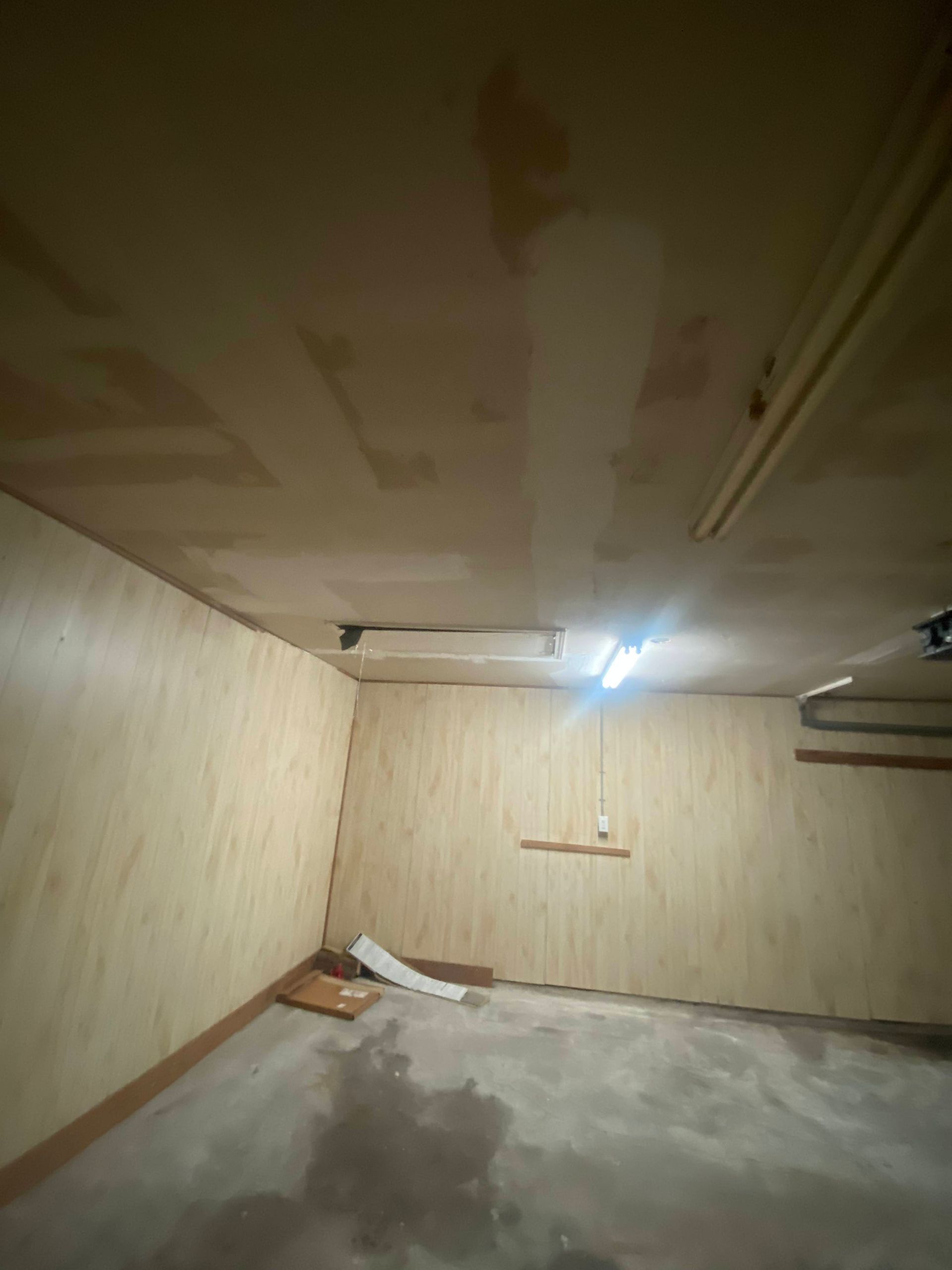 Empty room with plywood walls and ceiling, concrete floor. Fluorescent lights.