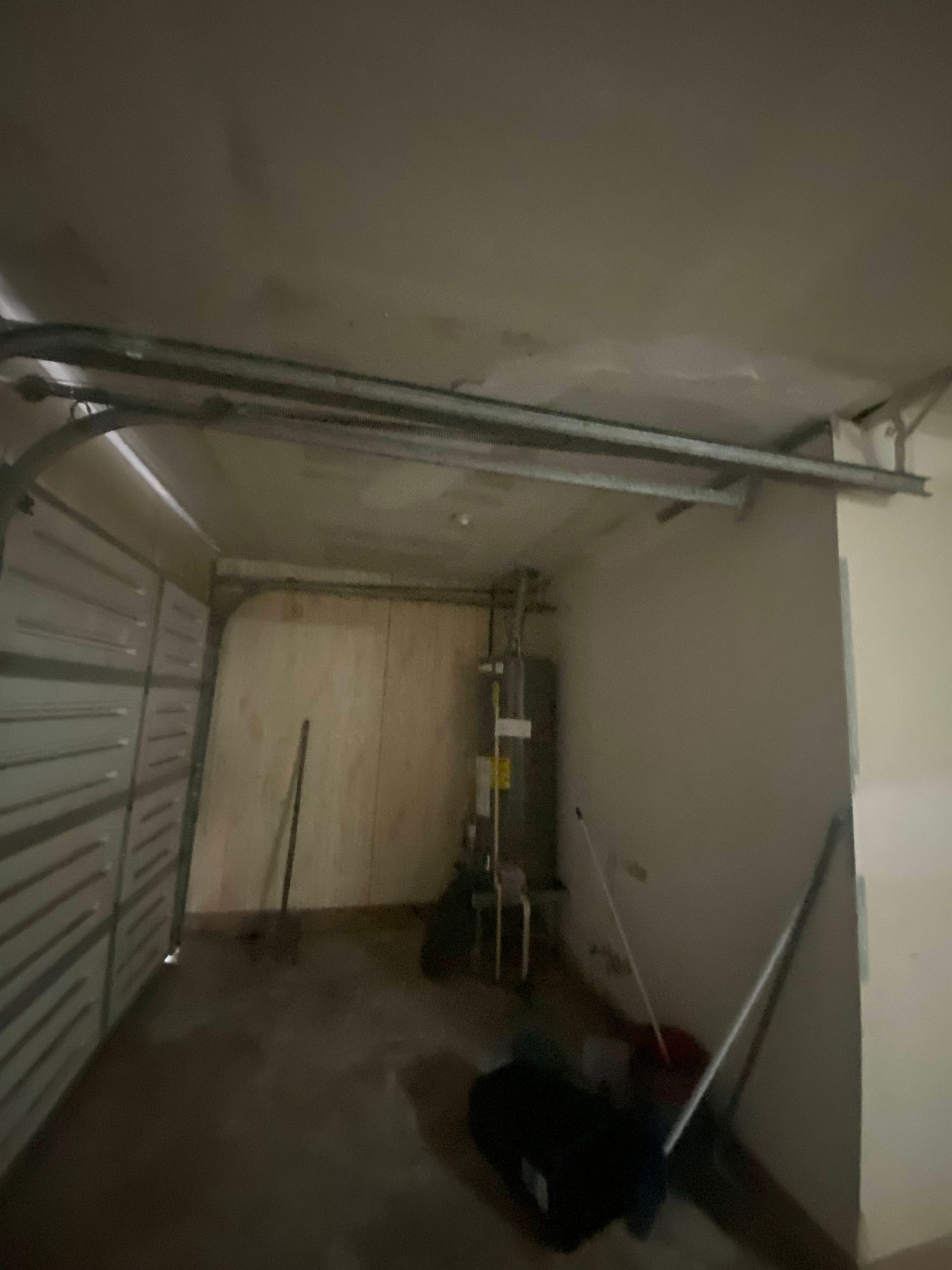 Interior of an empty garage with a water heater, garage door tracks, and various items leaning against the wall.