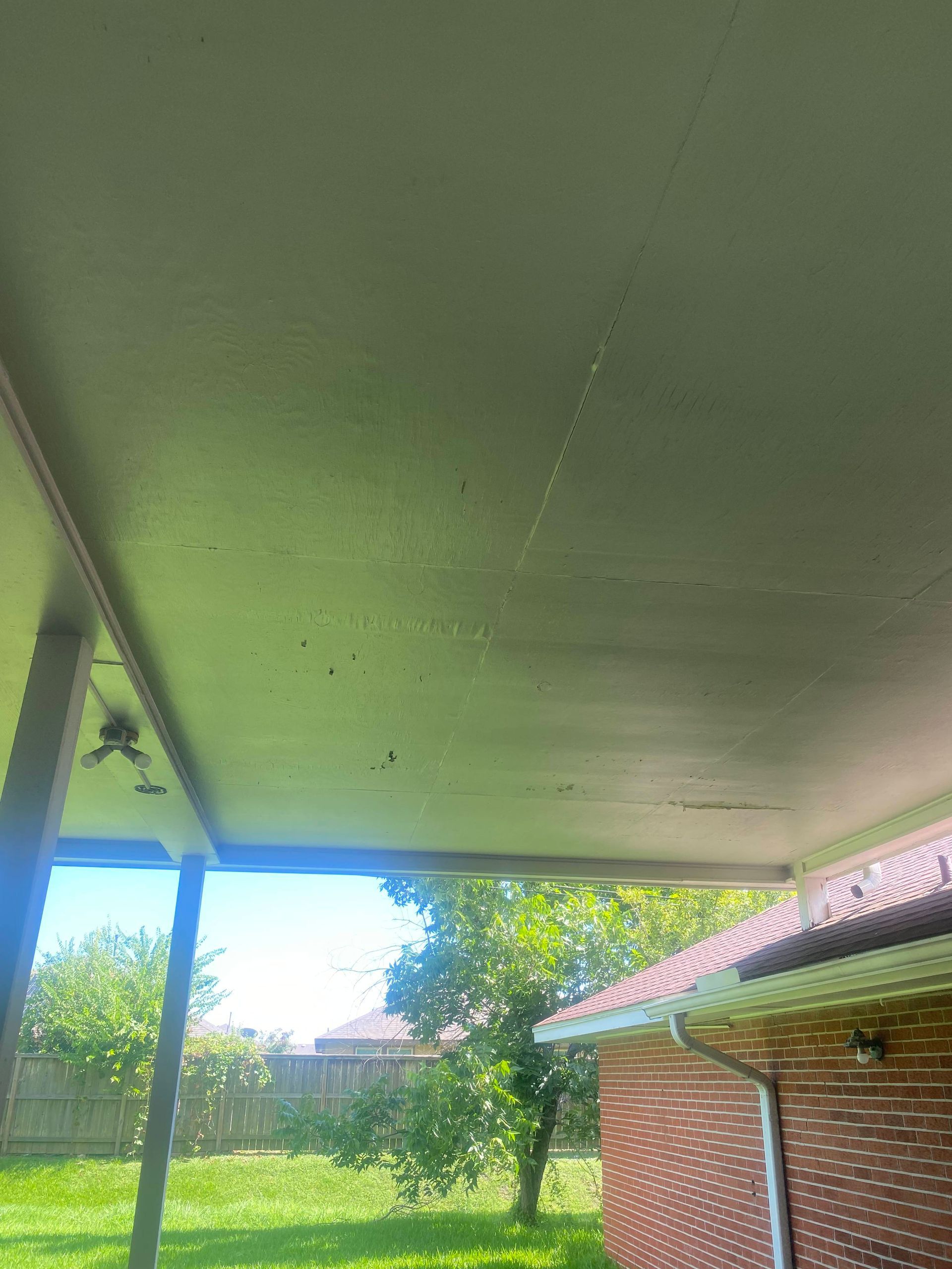 Overhead view of a covered patio ceiling, green-painted, with cracks, and a glimpse of the sunny outdoors.