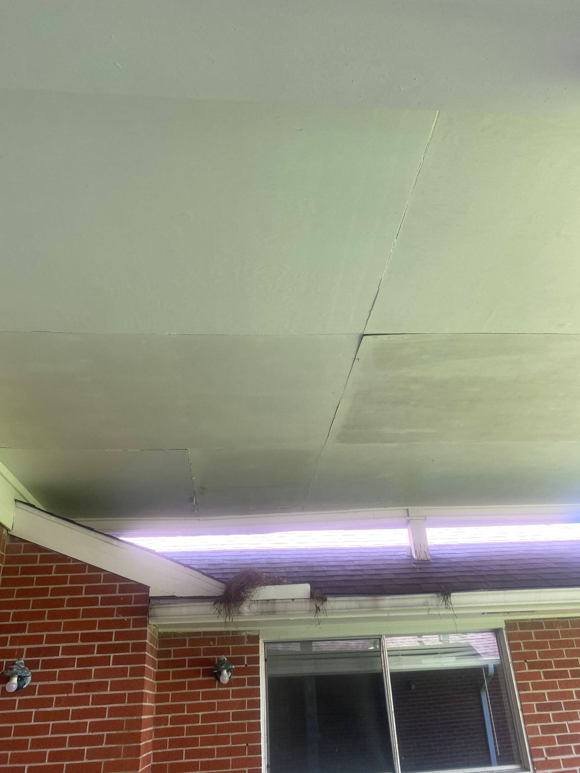 Ceiling of a porch with cracks, painted white. Brick wall and window frame visible below.