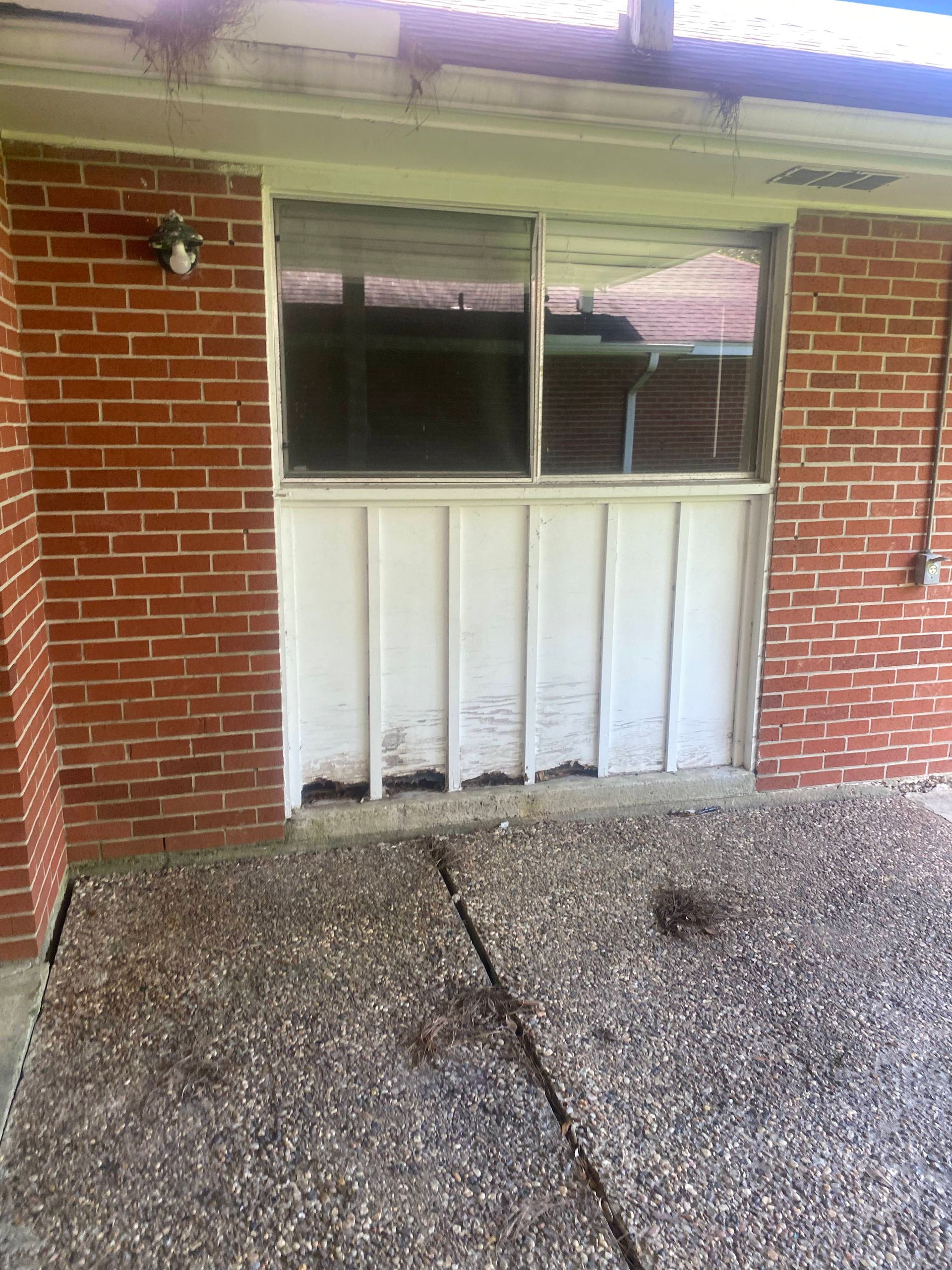 Brick wall with window above a damaged white panel. Concrete patio in foreground.