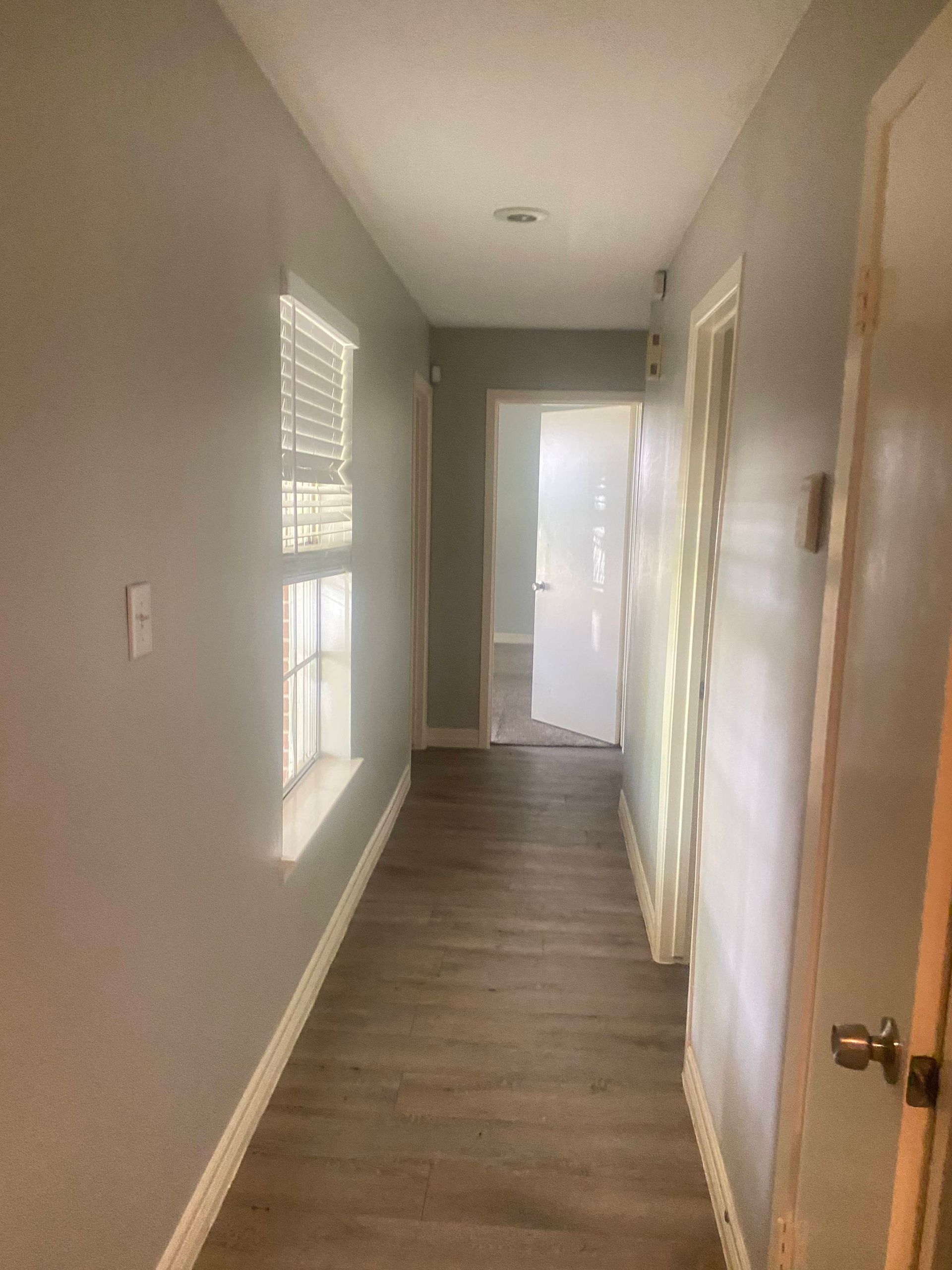 Narrow hallway with gray walls, wood-look flooring, and a window. Doorways on the right and at the end.