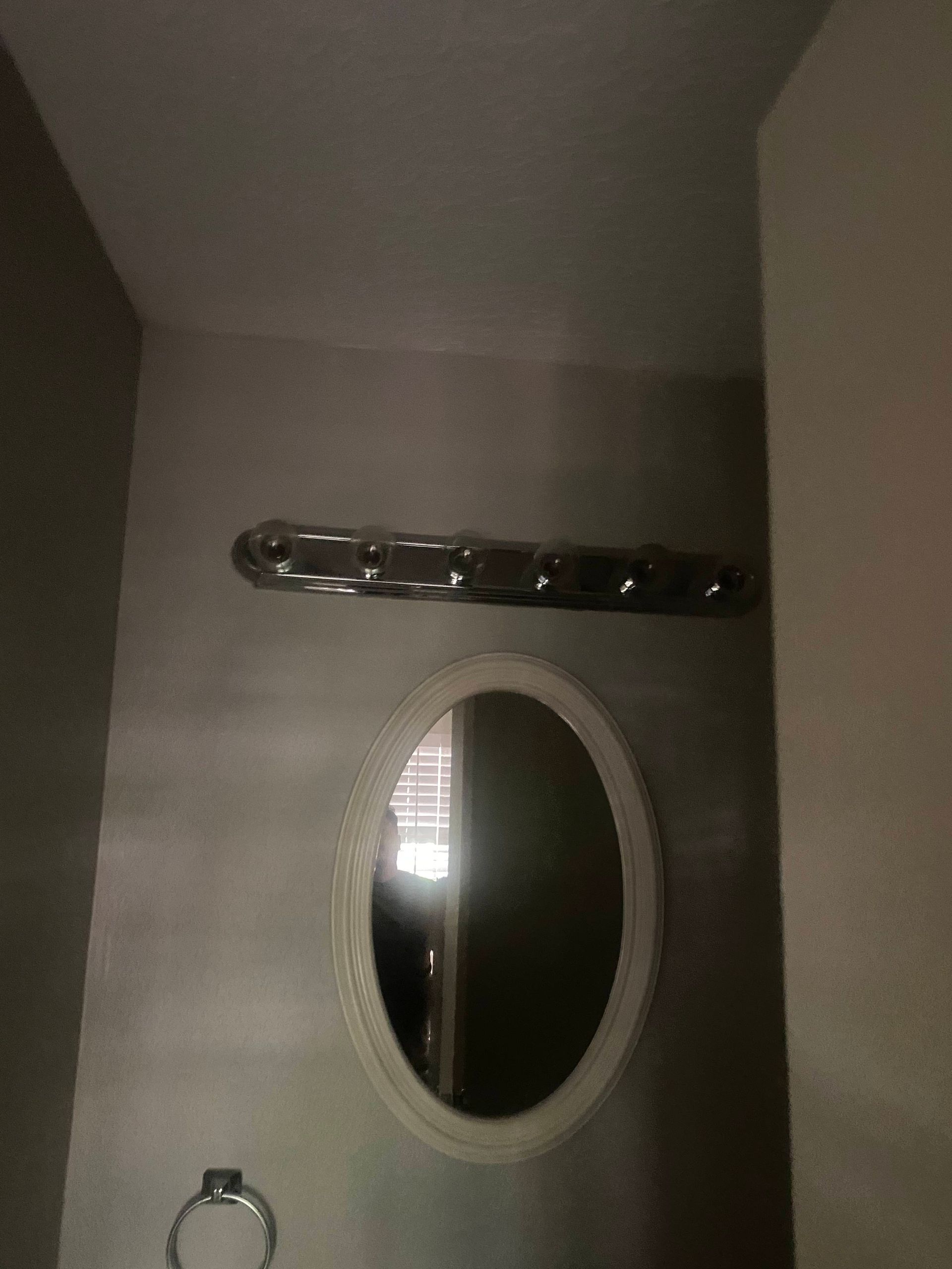 Oval mirror and light fixture in a bathroom. Beige wall and small towel ring.