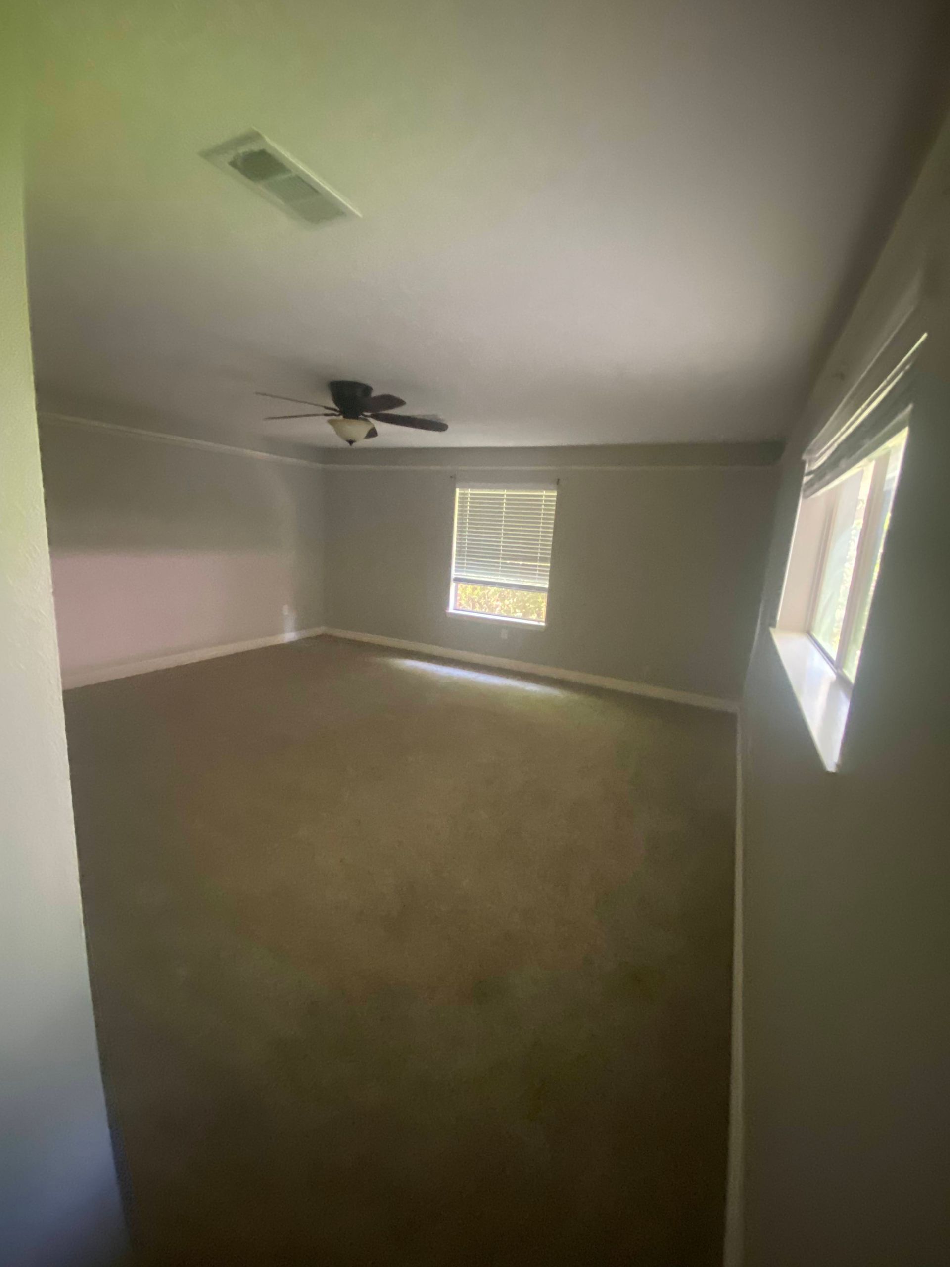 Empty room with brown carpet, gray walls, and ceiling fan. Window on right, air vent on left.
