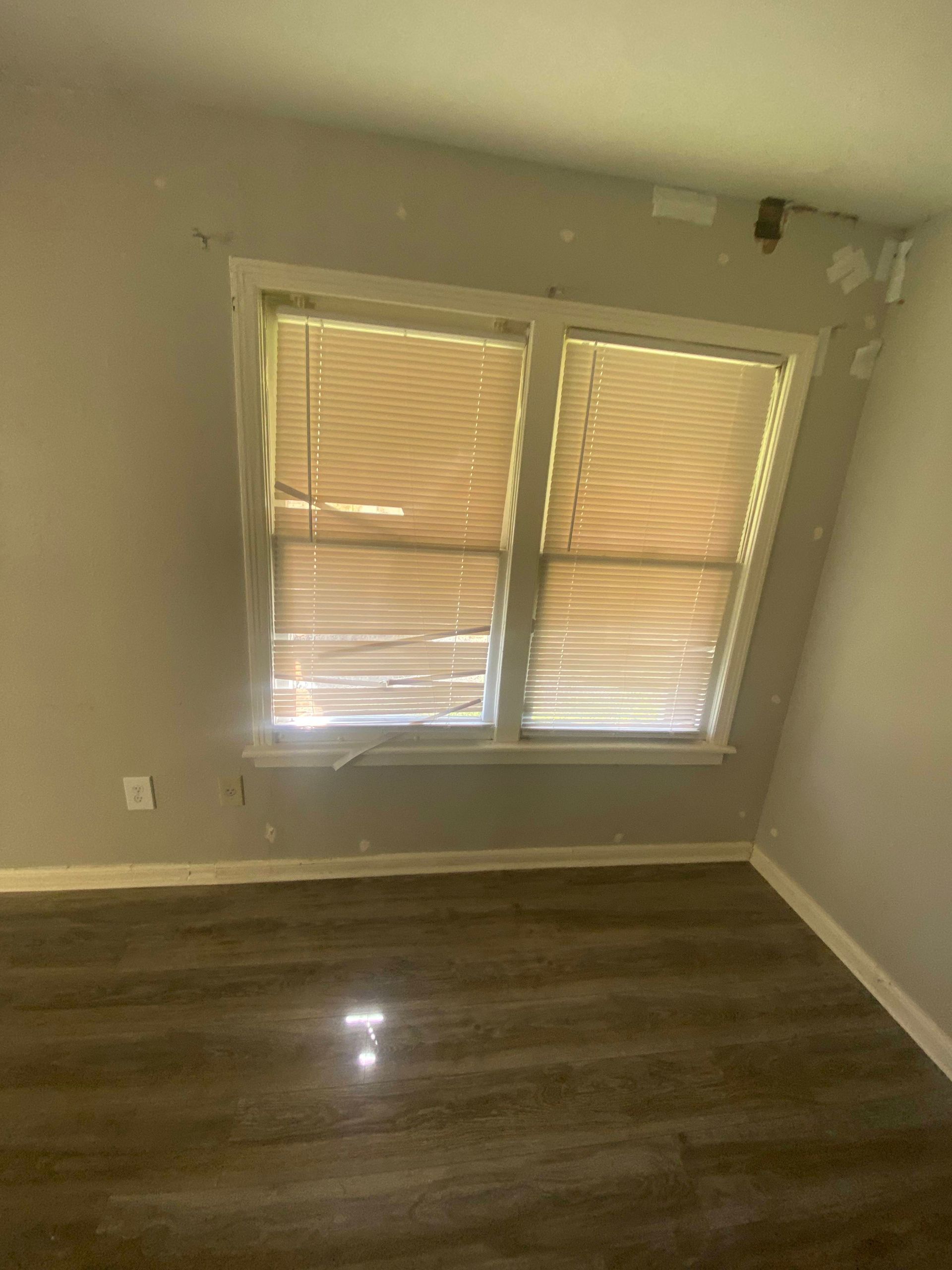 Empty room with a window, blinds closed. Grey walls, dark wood-look floor, sunlight visible.