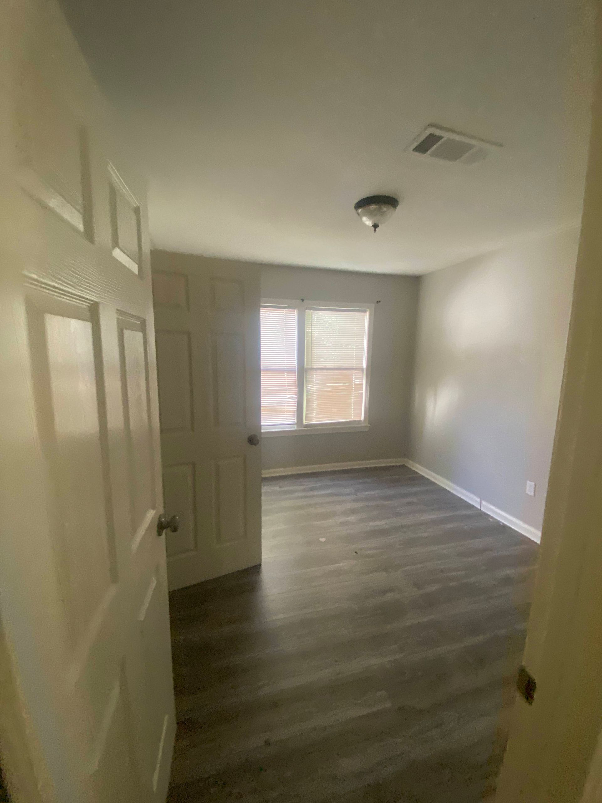 Empty room with gray wood-look flooring, a window with blinds, and a closed white door.