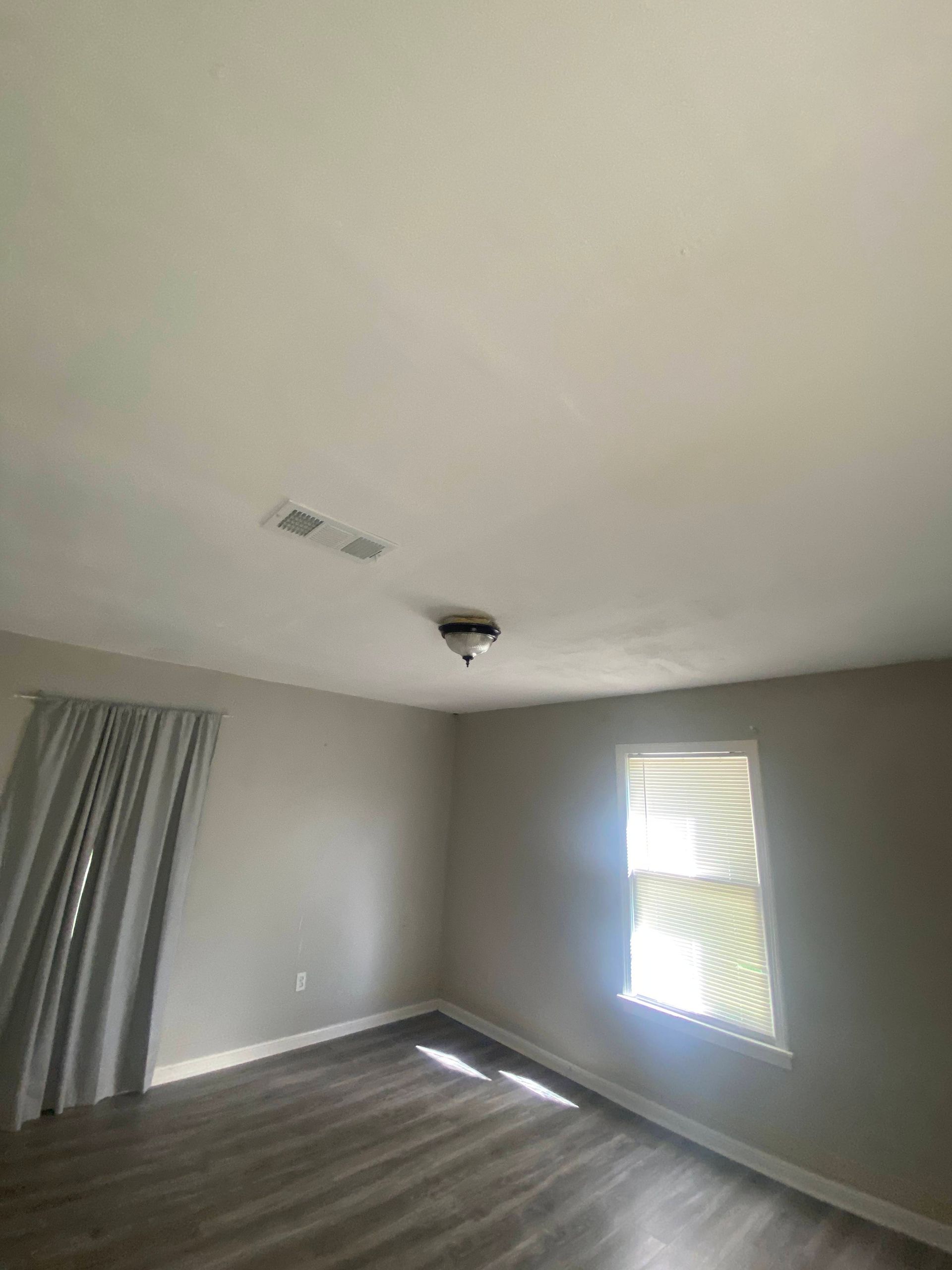 An empty room with grey walls and wood-look flooring. A window with light, grey curtains, and ceiling light fixture.