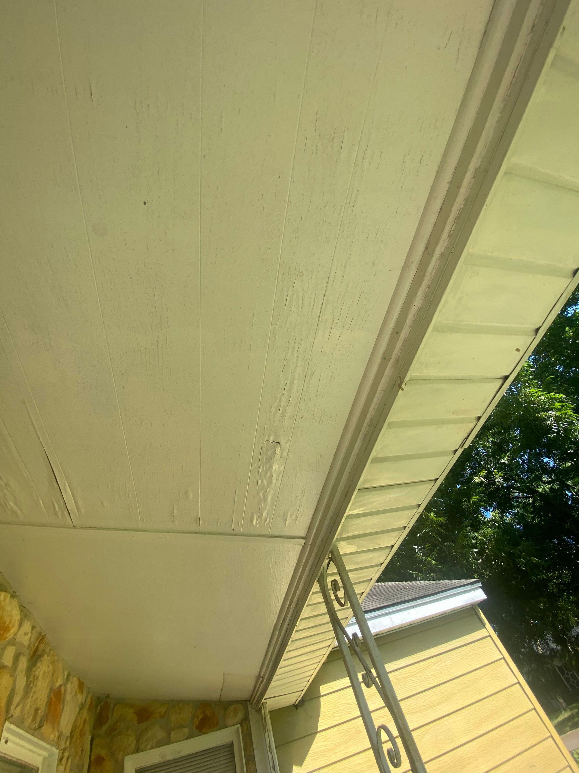 View of a porch ceiling, cream-colored vinyl siding with slight water stains. Sunlight brightens the scene.