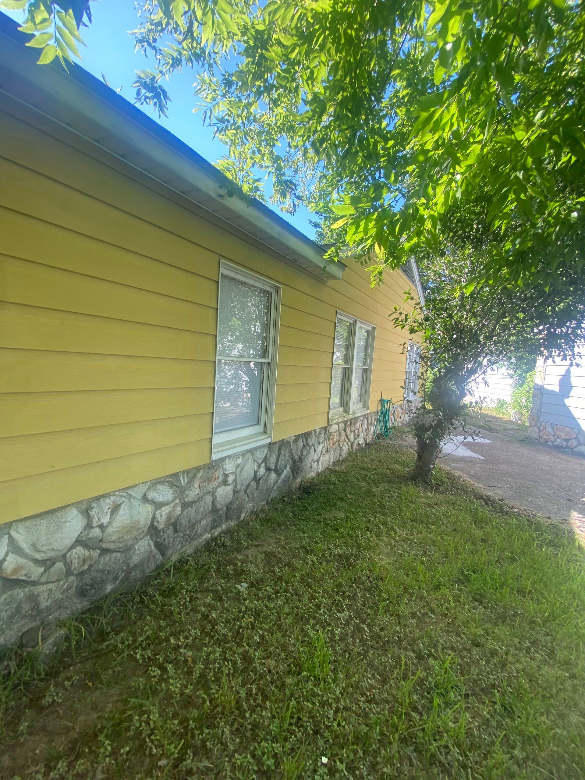 Yellow house with stone foundation and windows, shaded by a tree on a sunny day.