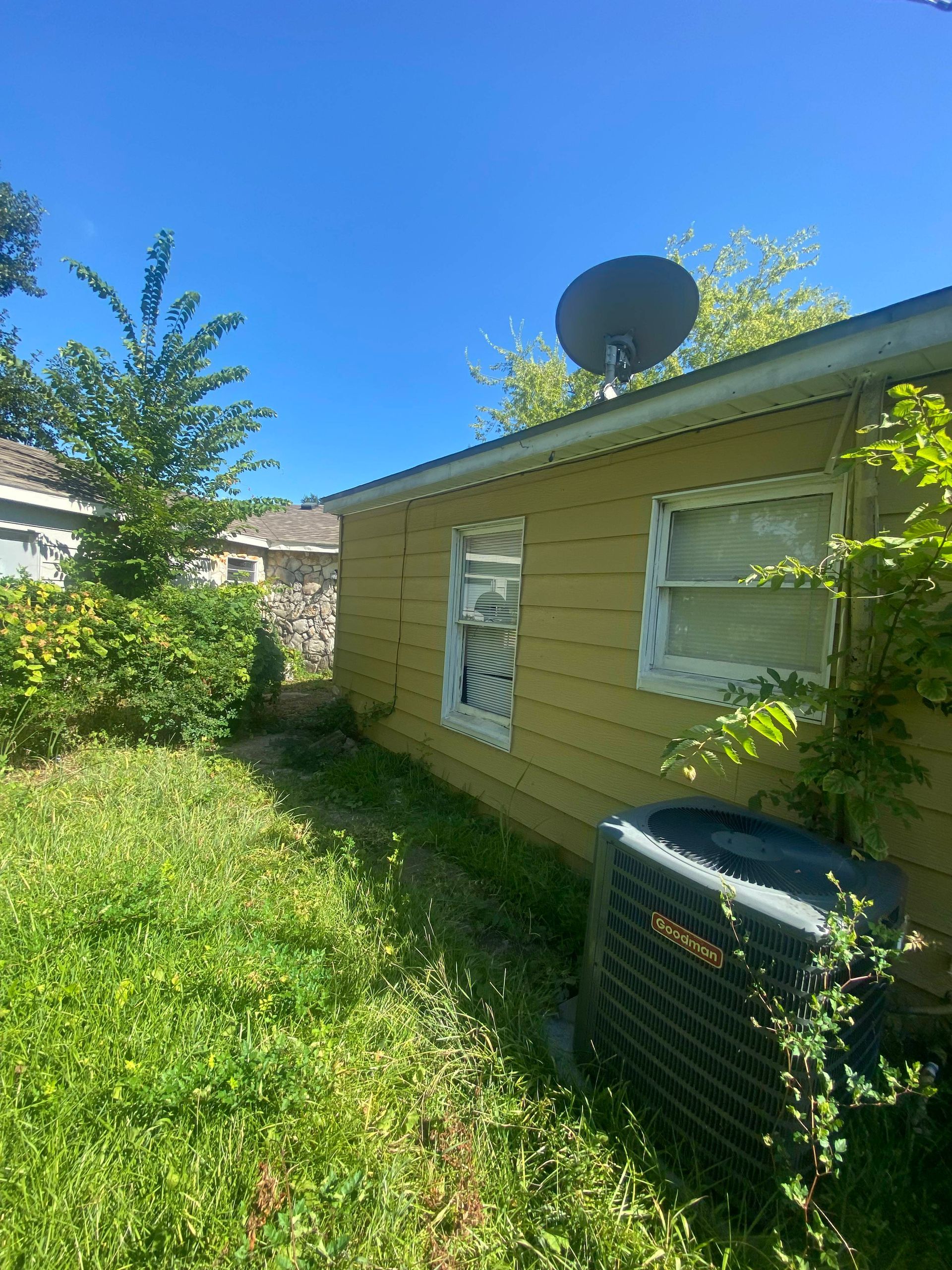 Yellow house exterior with satellite dish, air conditioner, and overgrown lawn under a blue sky.
