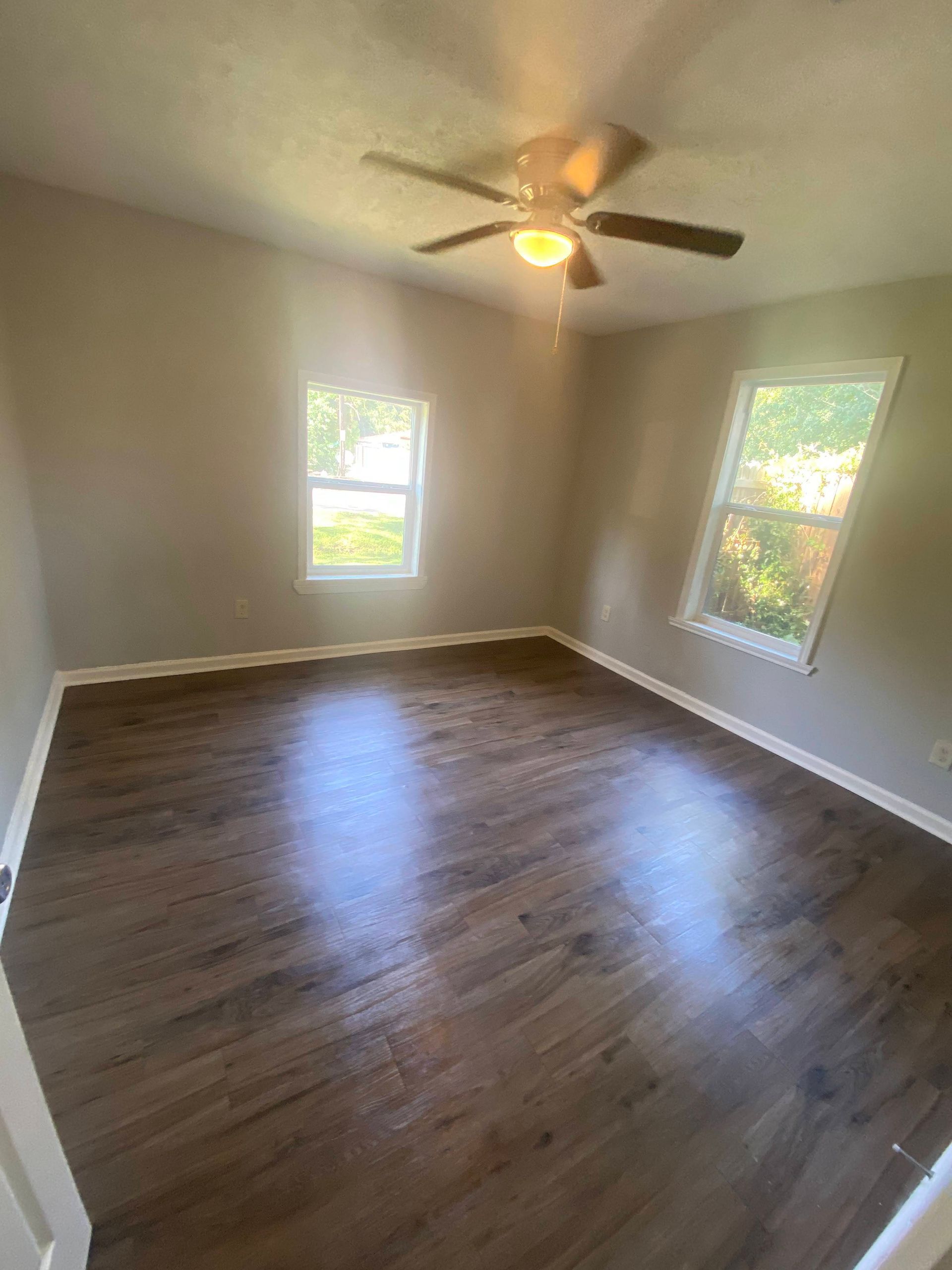 Empty room with dark wood-look flooring, two windows, beige walls, and a ceiling fan.