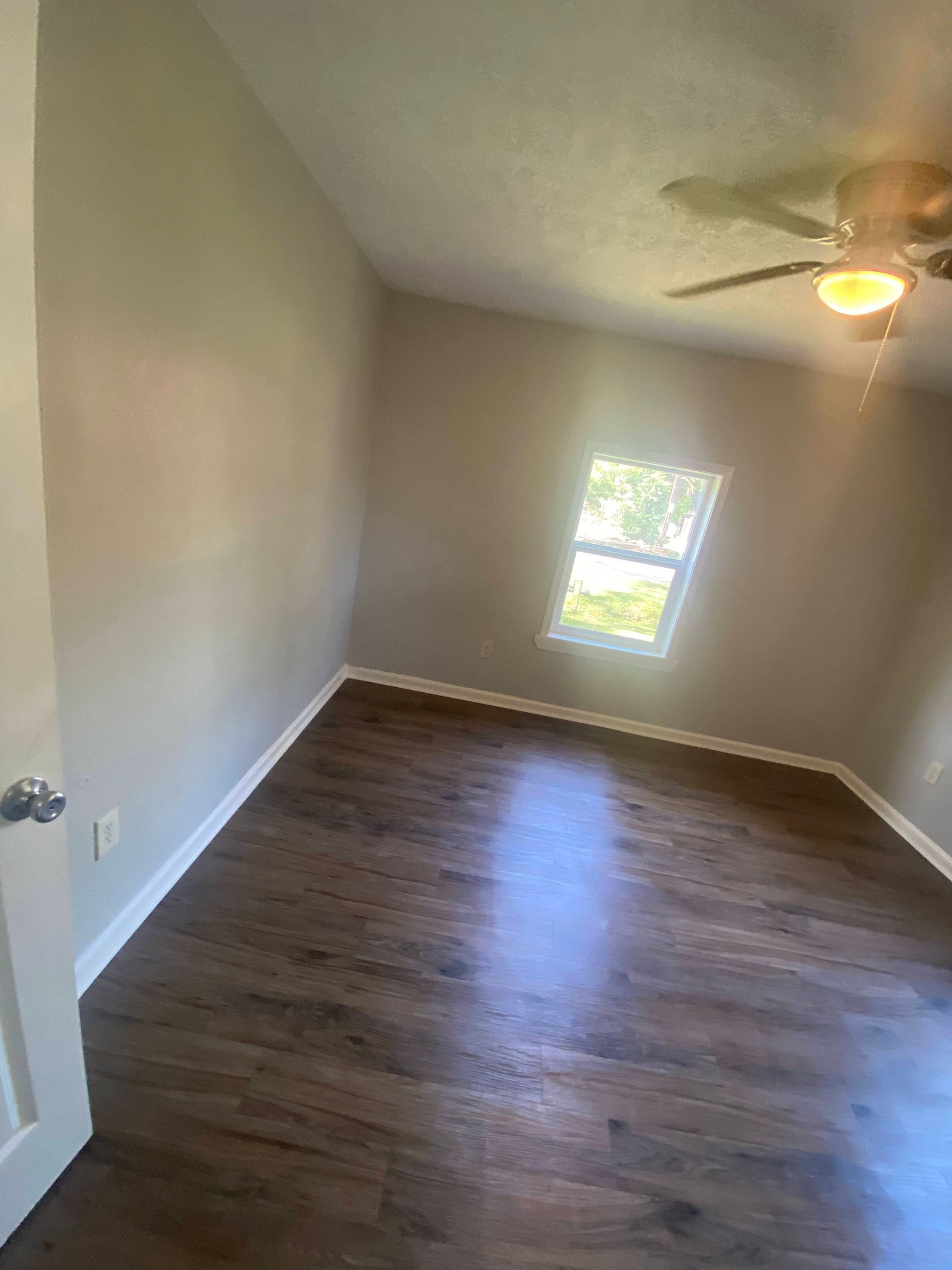 Empty room with brown flooring, tan walls, small window, and ceiling fan.