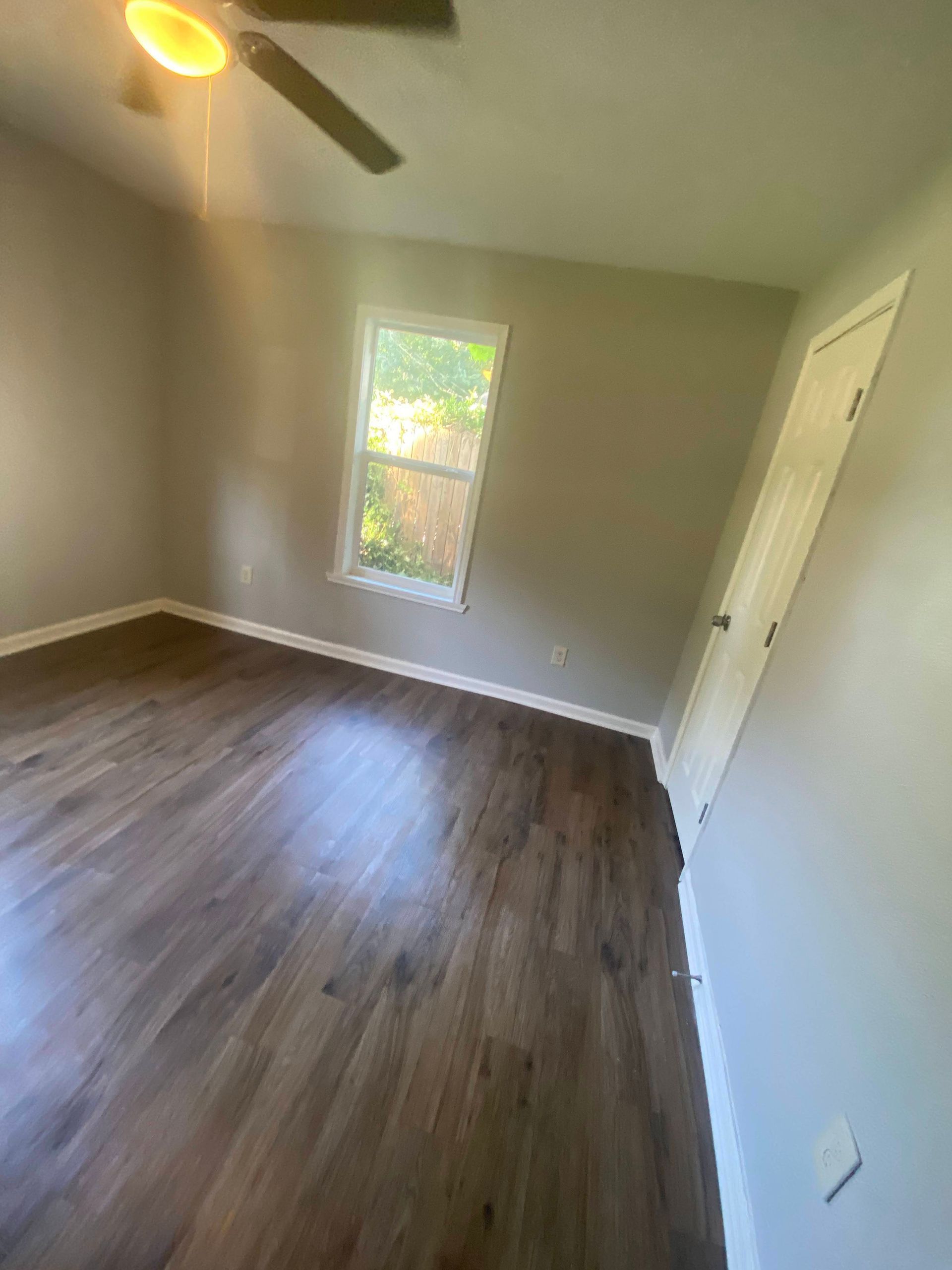 Empty room with dark wood-look flooring, gray walls, a window, a door, and a ceiling fan.