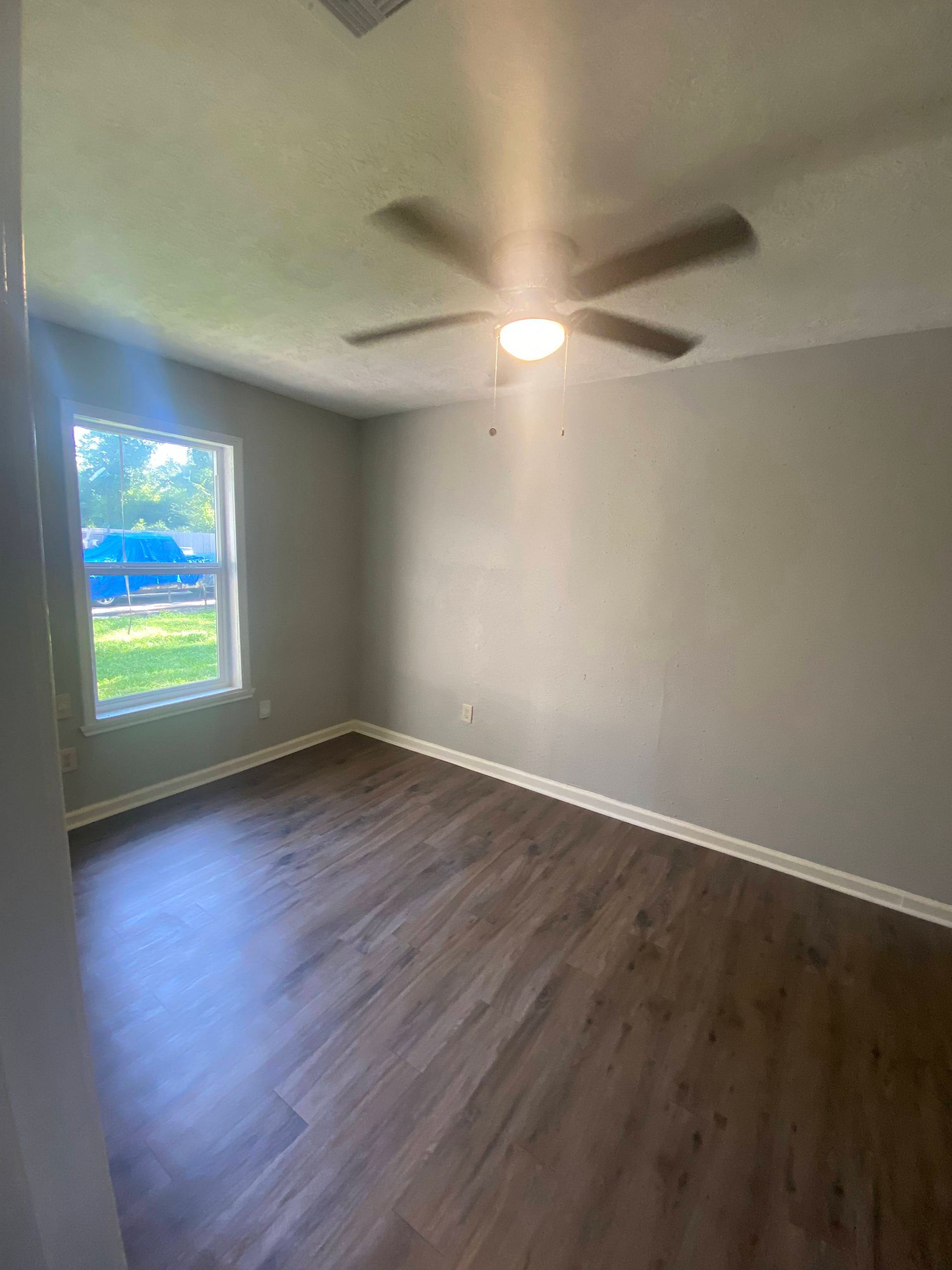 Empty room with gray walls, dark wood-look floor, and a ceiling fan. A window is in the left corner.