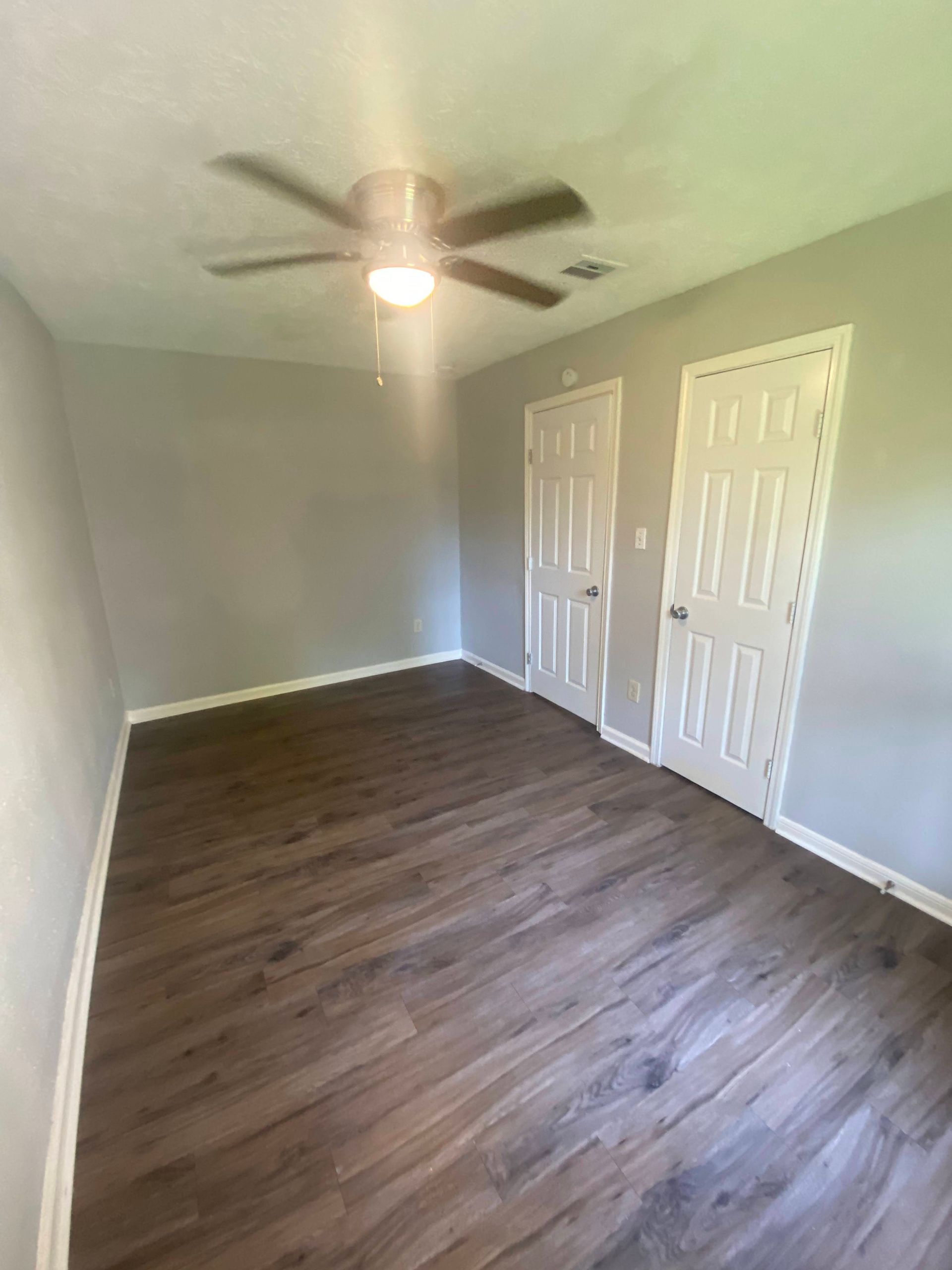 Empty room with gray walls, two white doors, wood-look flooring, and a ceiling fan.