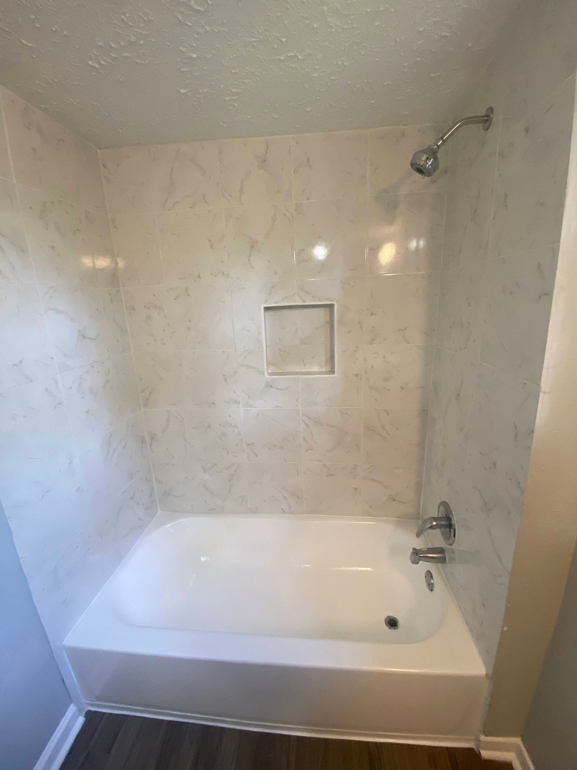 White tub and shower with light-colored faux marble walls and a built-in shelf.