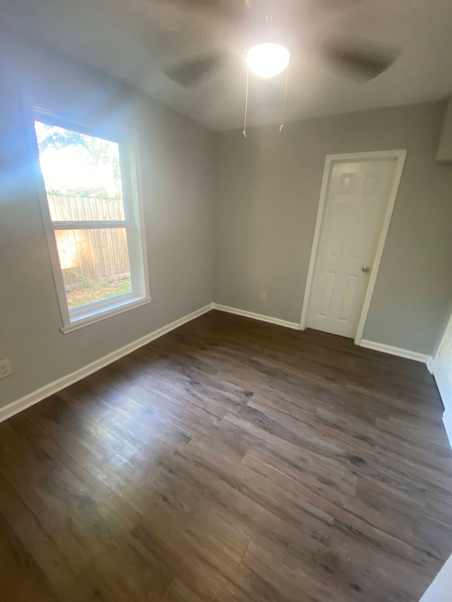 Empty bedroom with gray walls, wood-look flooring, white trim, and a window.
