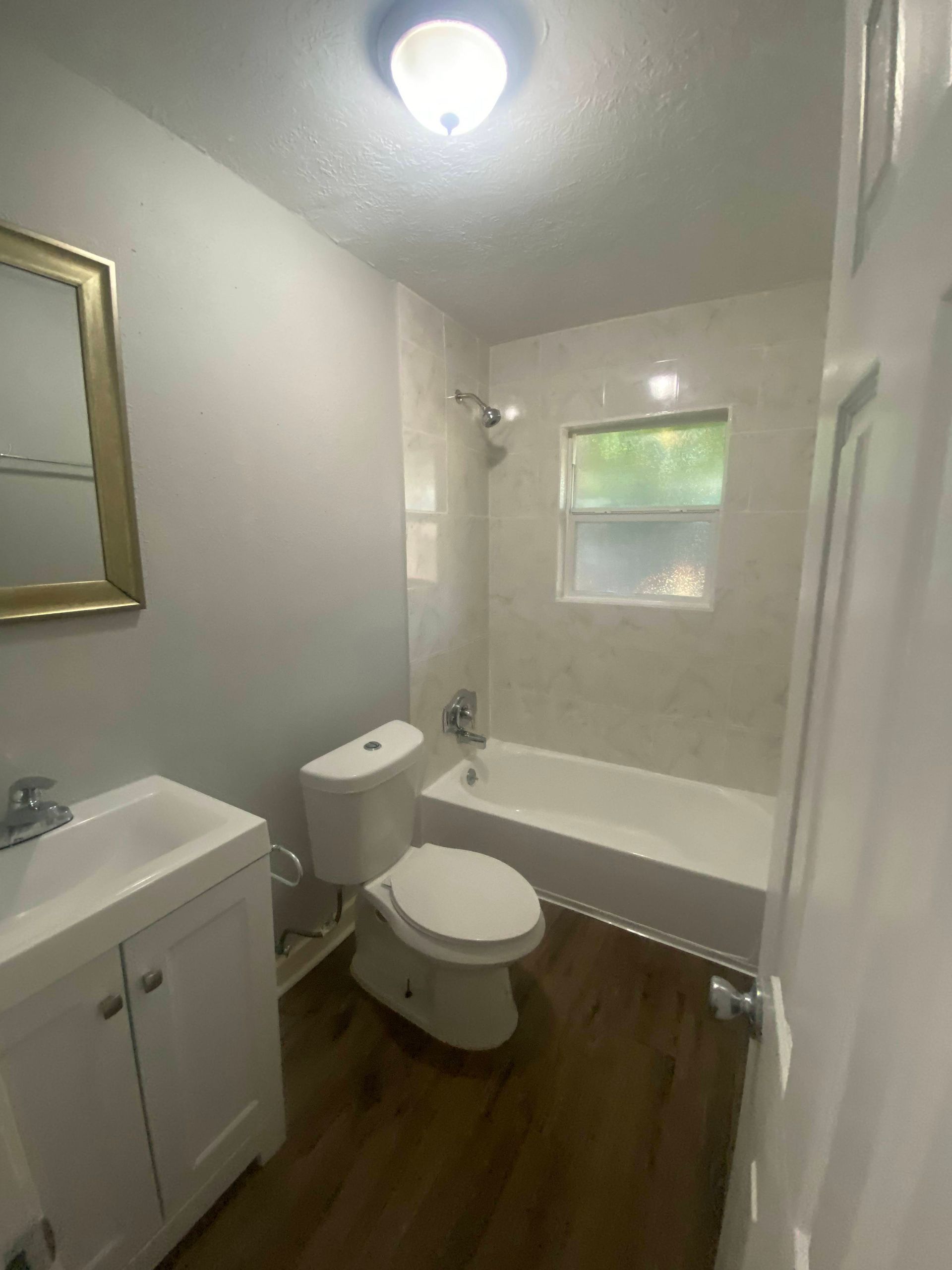 Bathroom with white vanity, toilet, and bathtub. Light gray walls, wood-look floor, and a bright overhead light.
