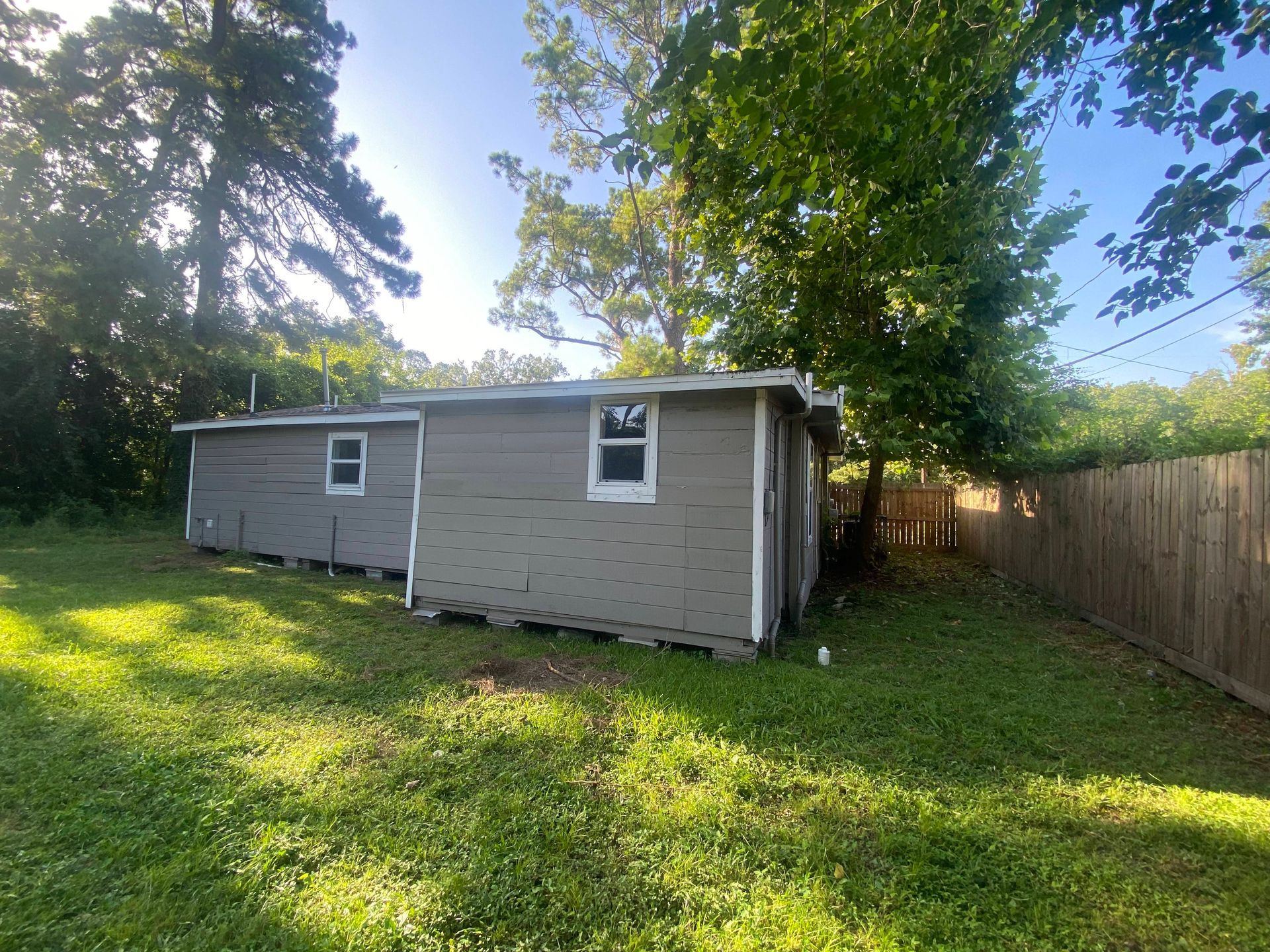 Gray building with two windows in a grassy yard, next to a wooden fence and trees.