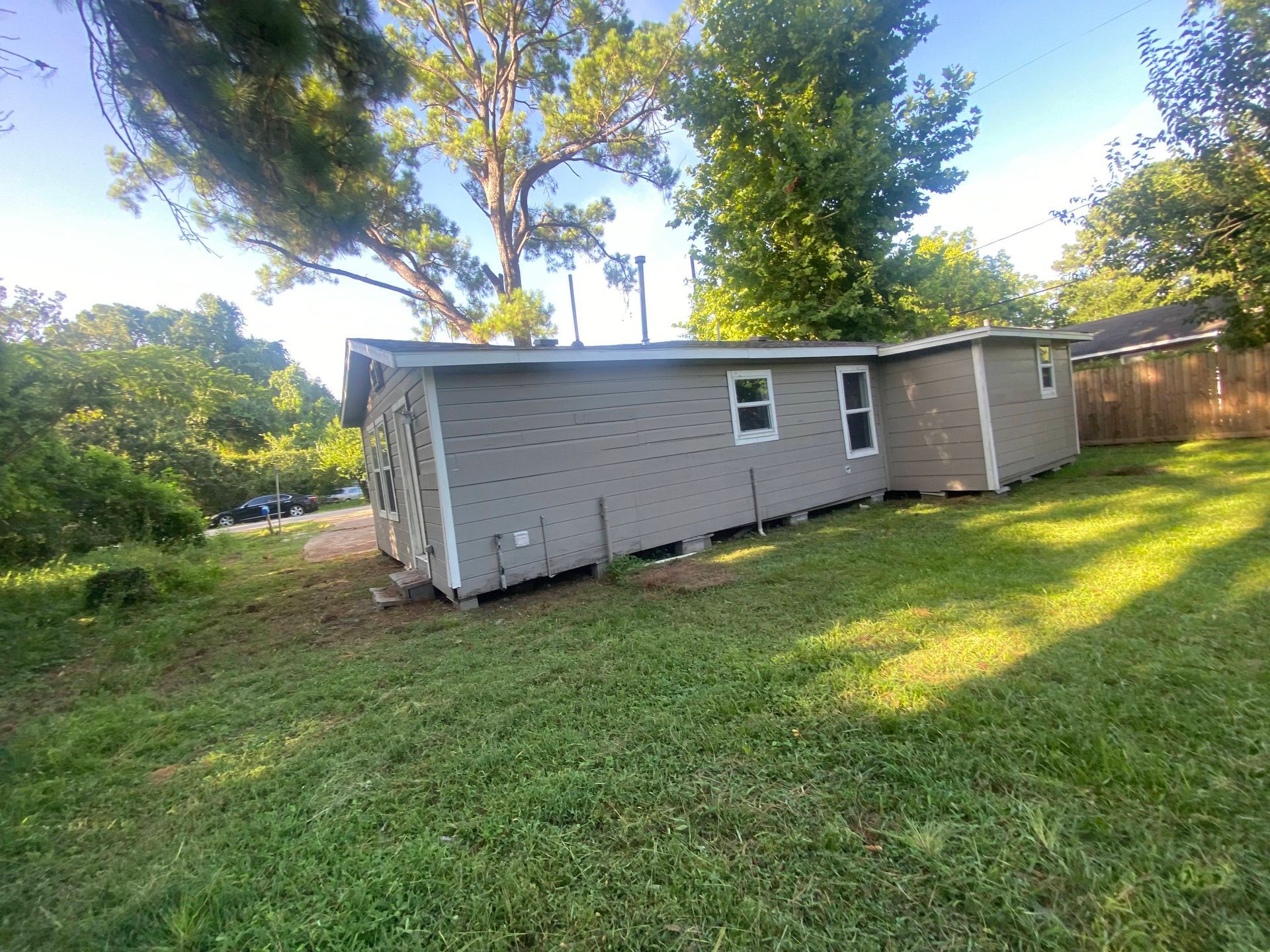 Gray house with white-framed windows on a grassy hill, surrounded by trees under a blue sky.