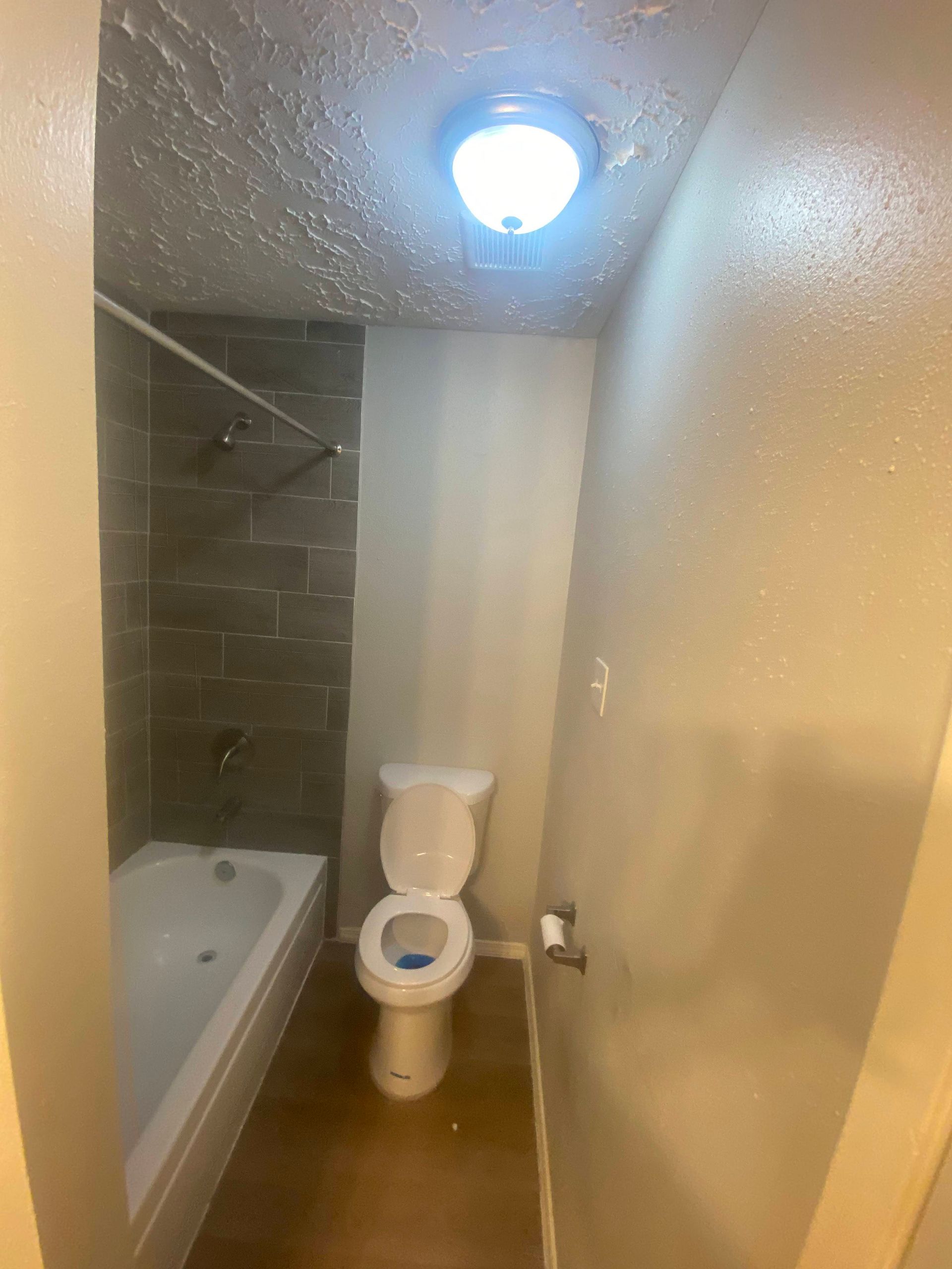 Small bathroom with tub, shower, and toilet; gray tile, white fixtures, light fixture on ceiling.
