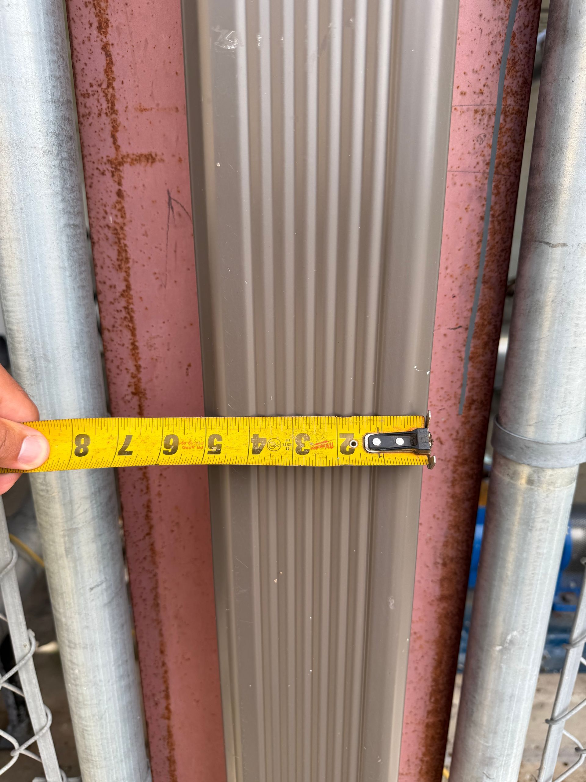 Hand measuring a brown gutter against a red fence and metal poles.