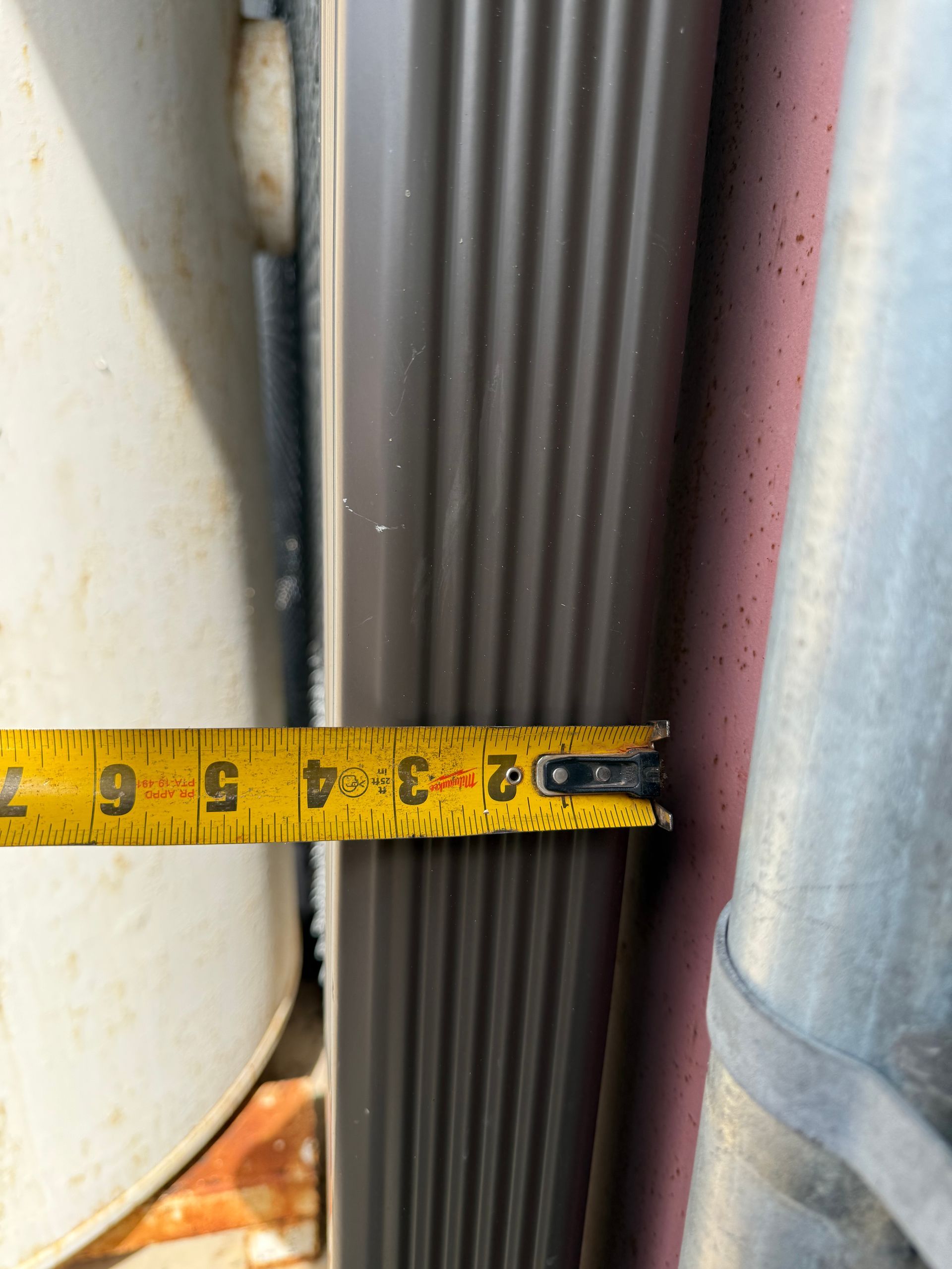 A tape measure shows approximately 3 inches between a brown corrugated vertical pipe and a round metal pipe.