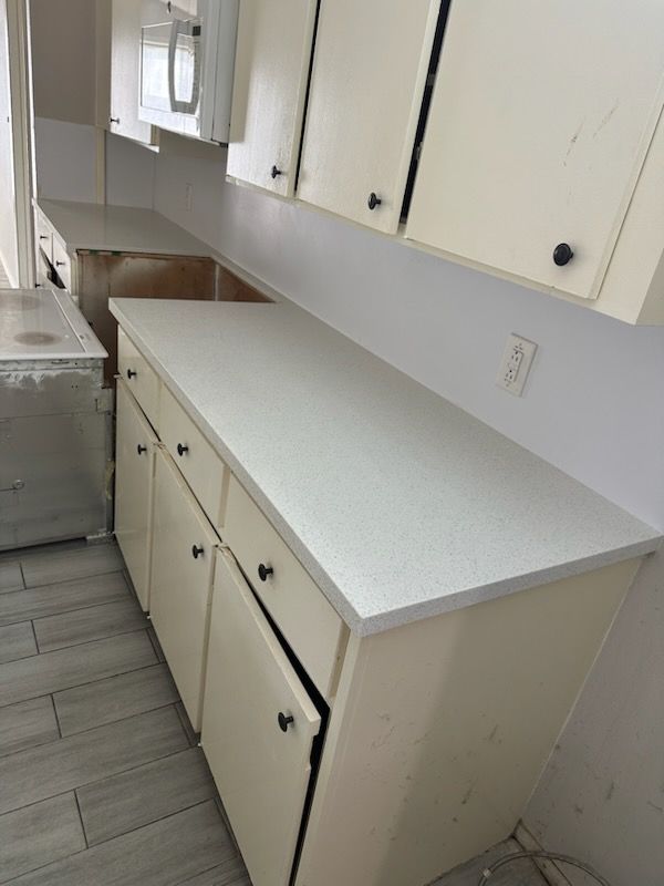 Cream-colored kitchen cabinets and countertop. Ajar cabinet door reveals interior. Beige walls, window and tiles.