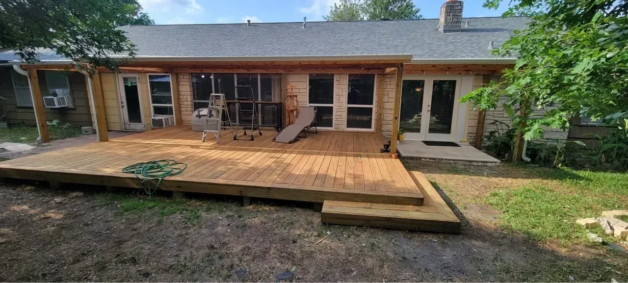 A wooden deck with steps leads to a house with multiple doors and windows. Green trees and grass are in the background.