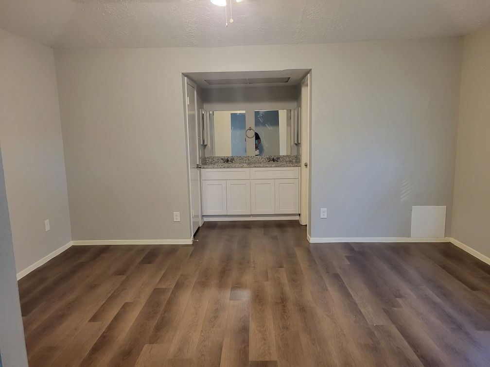 Empty room with wood floors and gray walls. A bathroom area is visible in the back.