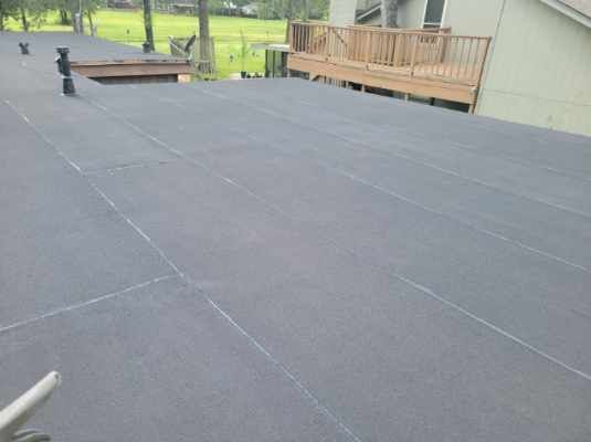 Flat, dark gray roof with visible seams. Wooden deck and house exterior in background.