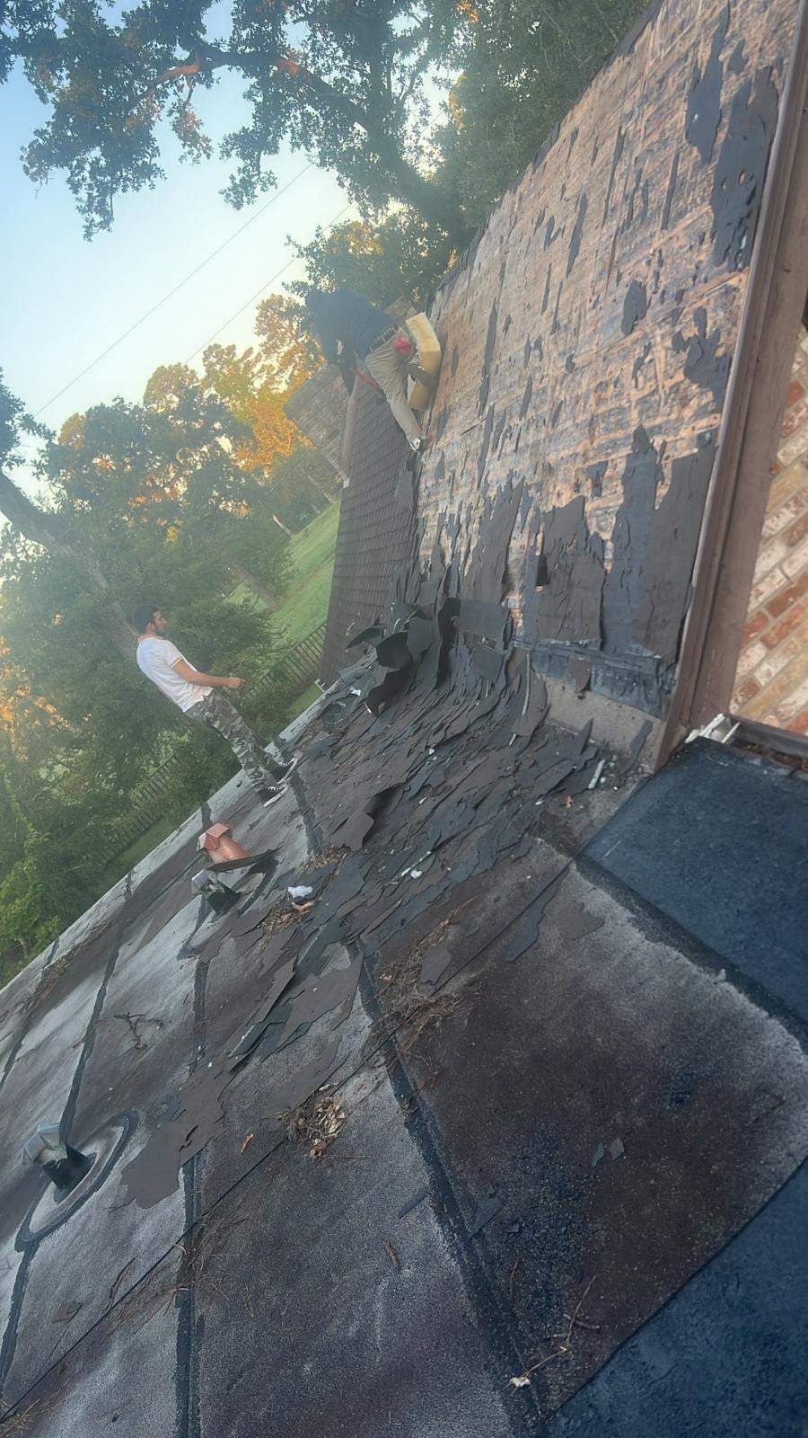 Person on roof near a brick chimney, appears to be working on the roof shingles. Trees and sky visible in the background.