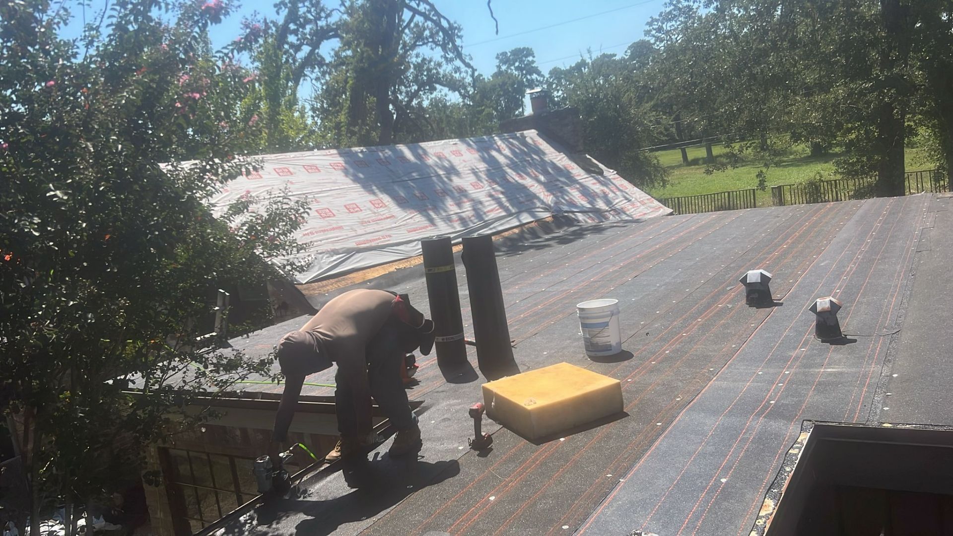 Roofer working on a flat roof. A partially completed roof with buckets, tools, and a sponge. Trees in the background.