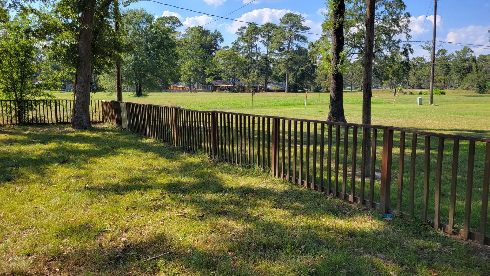 A brown wooden fence surrounds a grassy field with trees in the background under a blue sky.