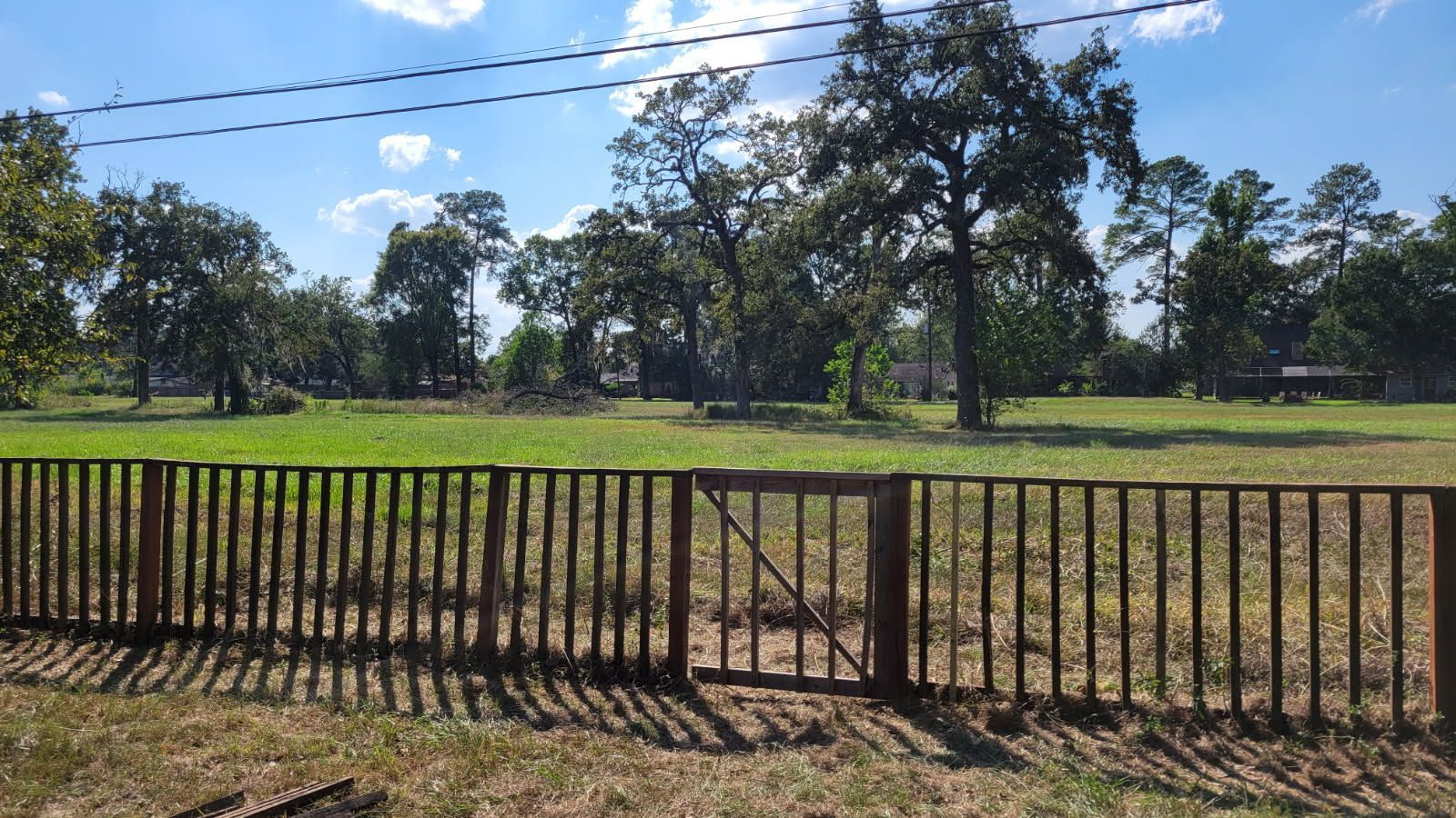 Wooden fence in front of a green field with trees under a blue sky.