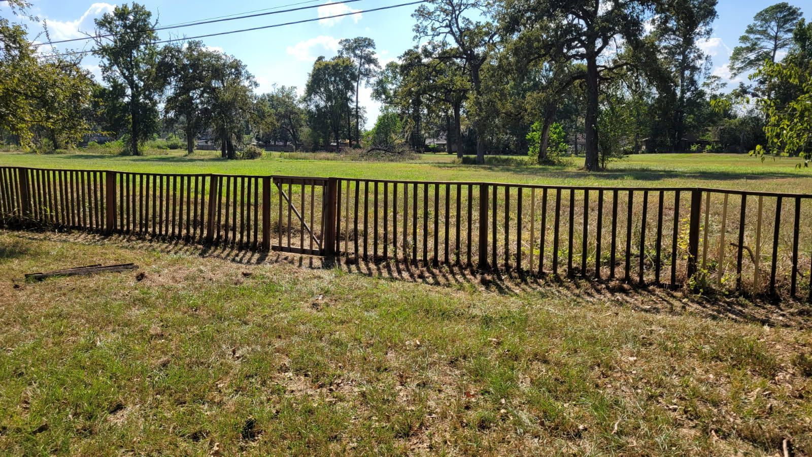 Wooden fence in grassy field with trees in the background under a blue sky.