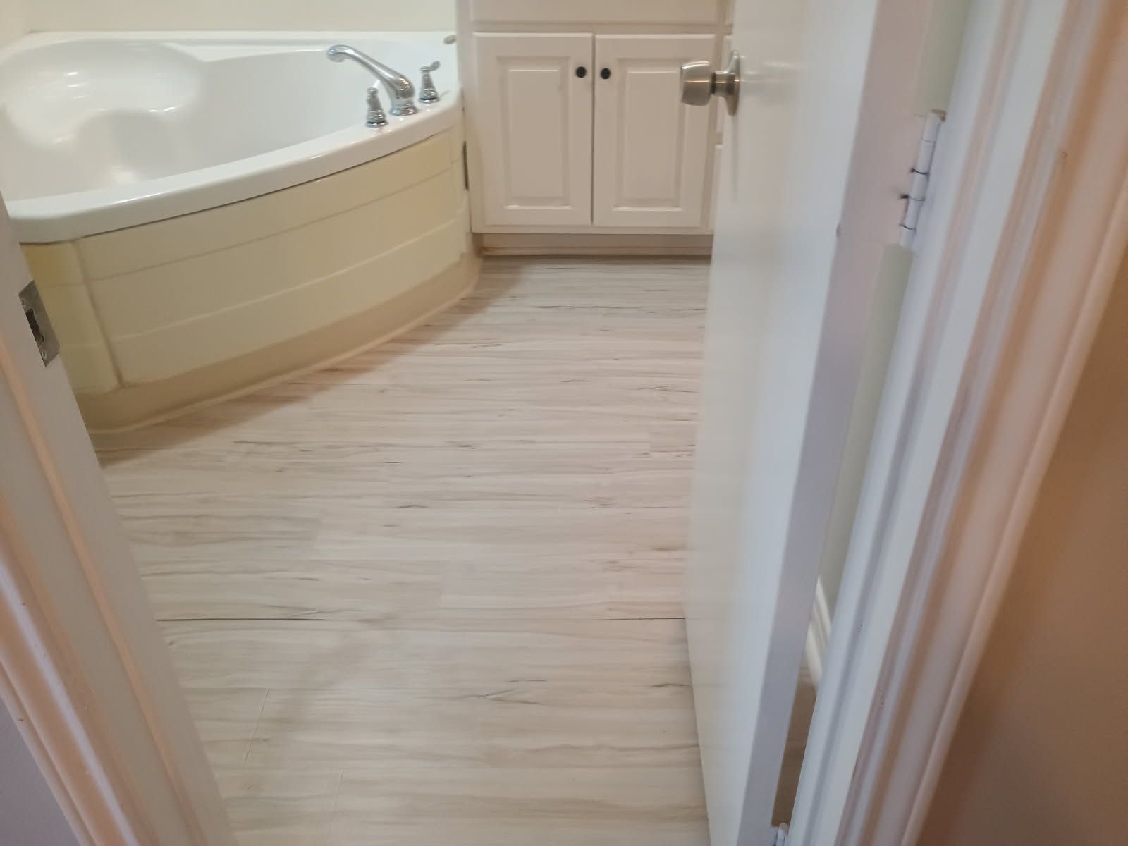 Bathroom with a white bathtub and wood-look flooring seen through a partially open door.