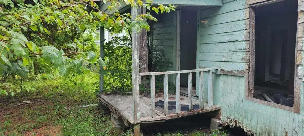 Dilapidated, blue-painted porch of an abandoned building overgrown with foliage.